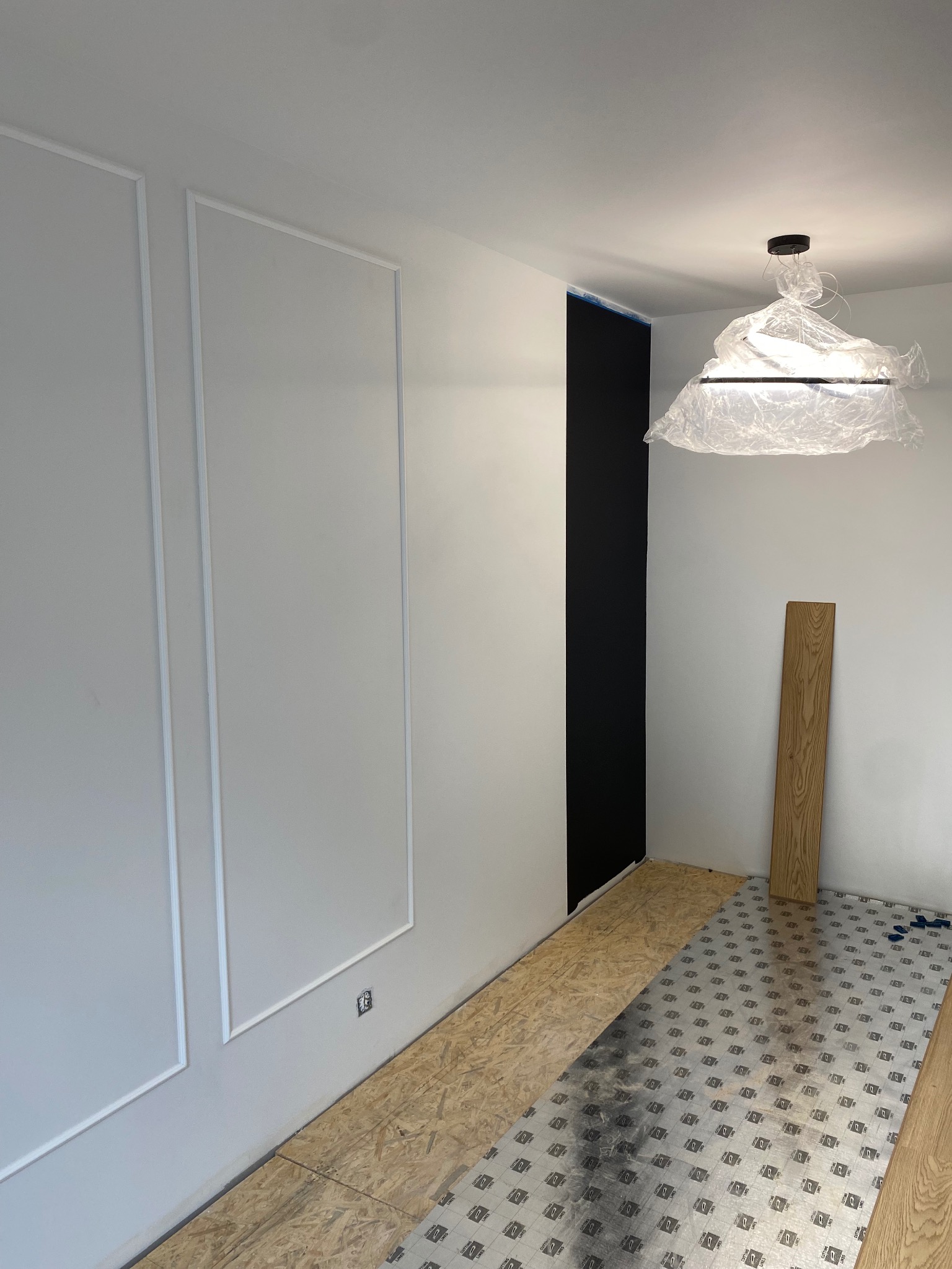 Wnętrze w trakcie prac wykończeniowych: białe ściany z dekoracyjnymi listwami, fragment czarnej ściany, podłoga przygotowana pod panele i lampa zabezpieczona folią