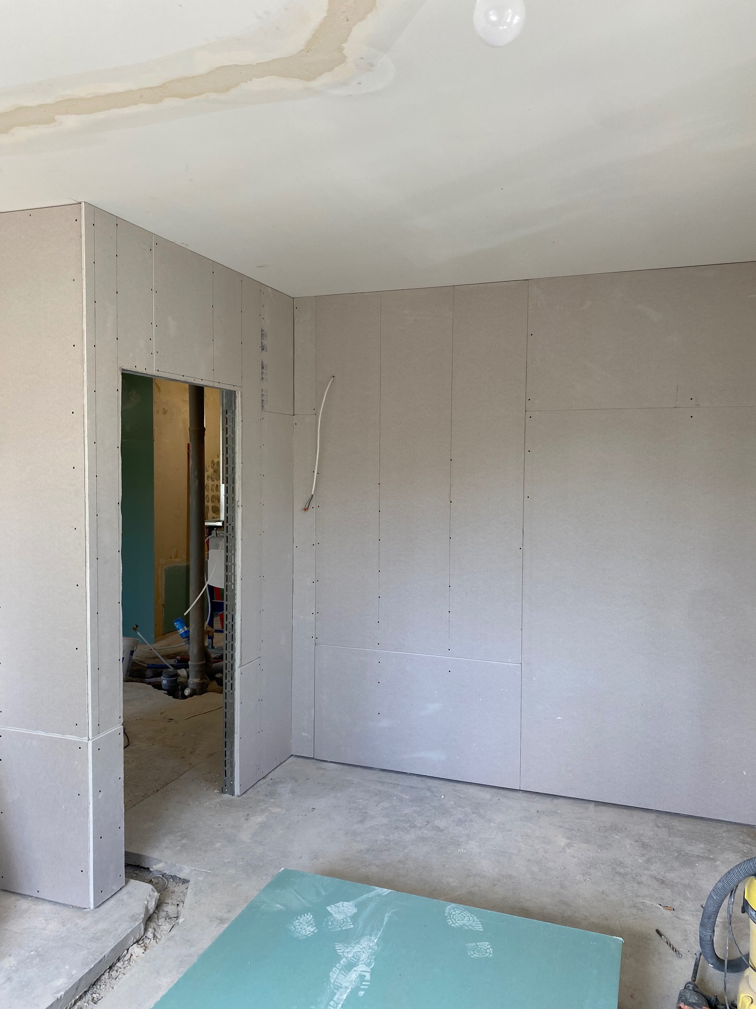 Montaż płyt karton gips na ścianach w trakcie remontu, widoczne ślady po zalaniu na suficie, niezagruntowana płyta na podłodze i otwór drzwiowy z widoczną instalacją.