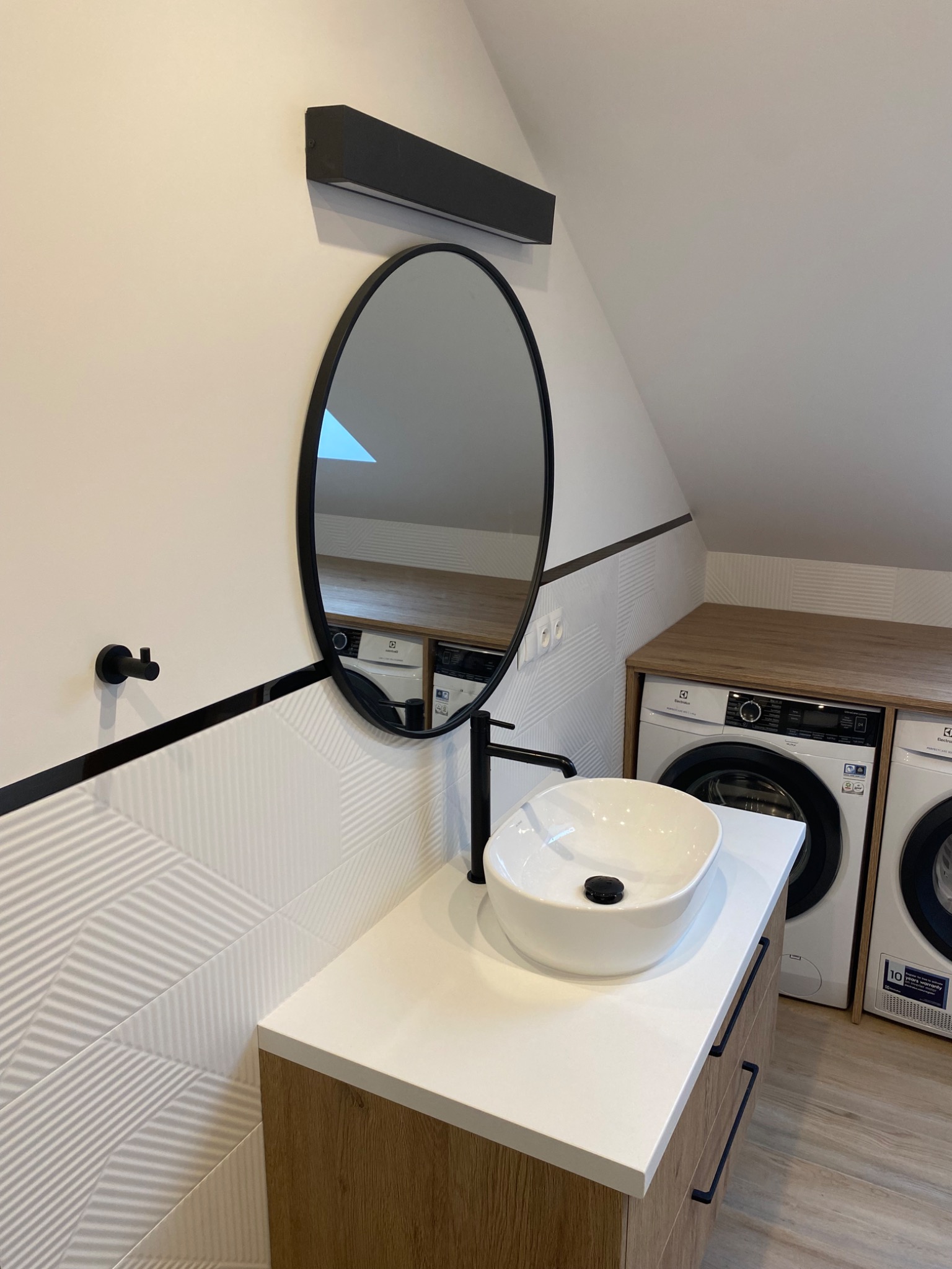 Nowoczesna łazienka na poddaszu z owalnym lustrem w czarnej ramie, białą umywalką nablatową z czarną armaturą, biało-szarą mozaiką na ścianie, zabudową pralki i suszarki z blatem w kolorze drewna.