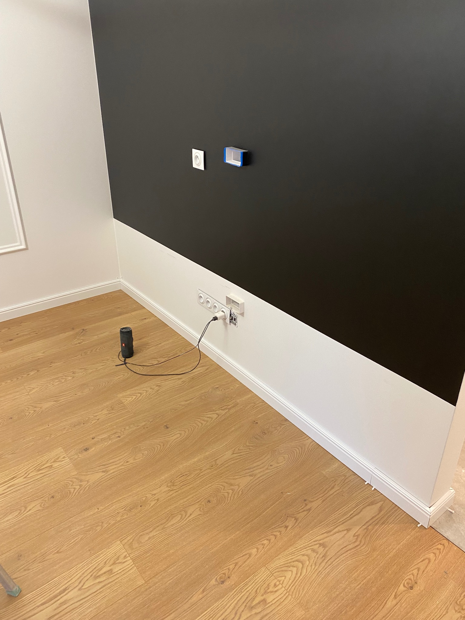 Pokój po malowaniu z dwukolorową ścianą: czarny pas na górze i biały na dole, drewniana podłoga, głośnik JBL podłączony do gniazdka.