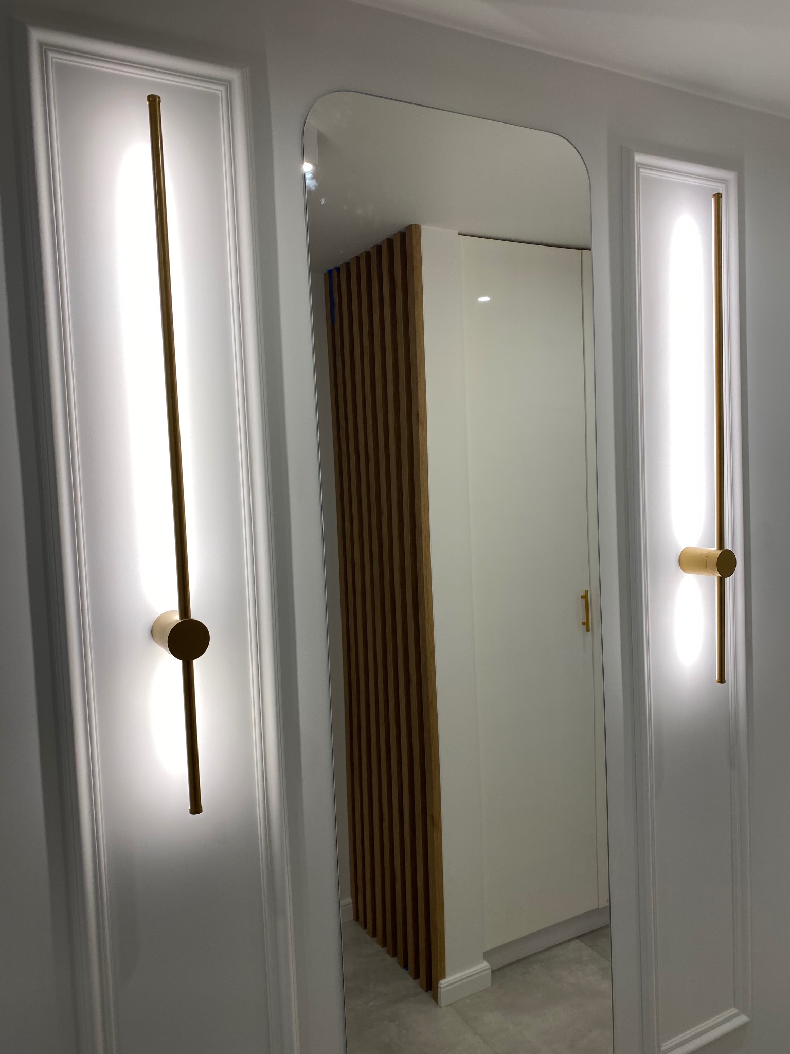 Nowoczesne oświetlenie ścienne zintegrowane z panelami dekoracyjnymi, widoczne odbicie szafy z drewnianymi akcentami w lustrze, minimalistyczny design wnętrza.