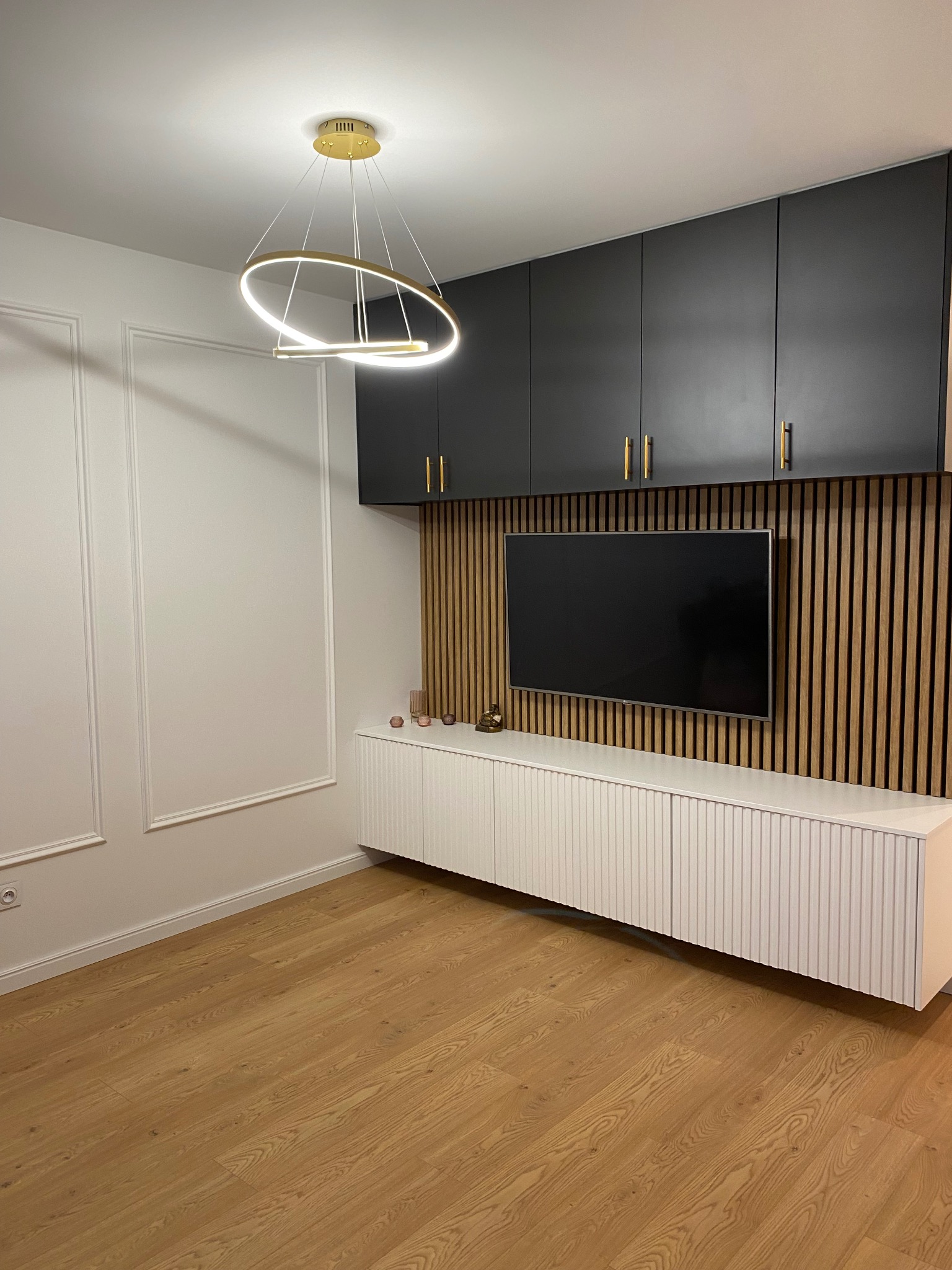 Nowoczesny salon z drewnianą ścianą z lamelami, wiszącą szafką z czarnymi frontami i złotymi uchwytami, białą szafką RTV z ryflowanymi drzwiczkami oraz okrągłą lampą wiszącą z elementami w kolorze...