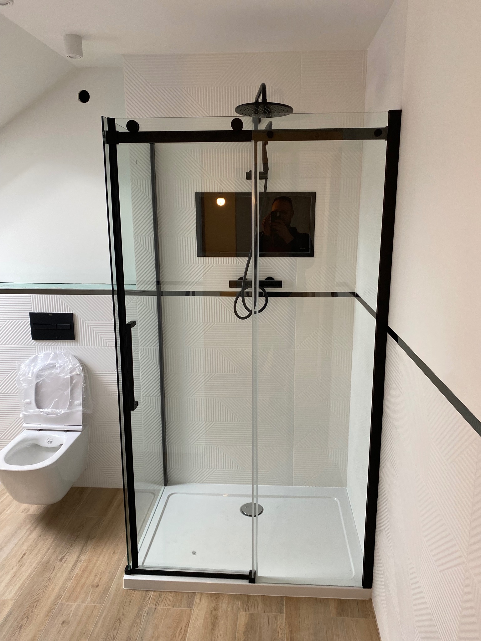 Nowoczesna łazienka w bieli i czerni z kabiną prysznicową typu walk-in z czarnymi profilami, wiszącą toaletą z osłoną ochronną i płytkami ściennymi o geometrycznym wzorze.