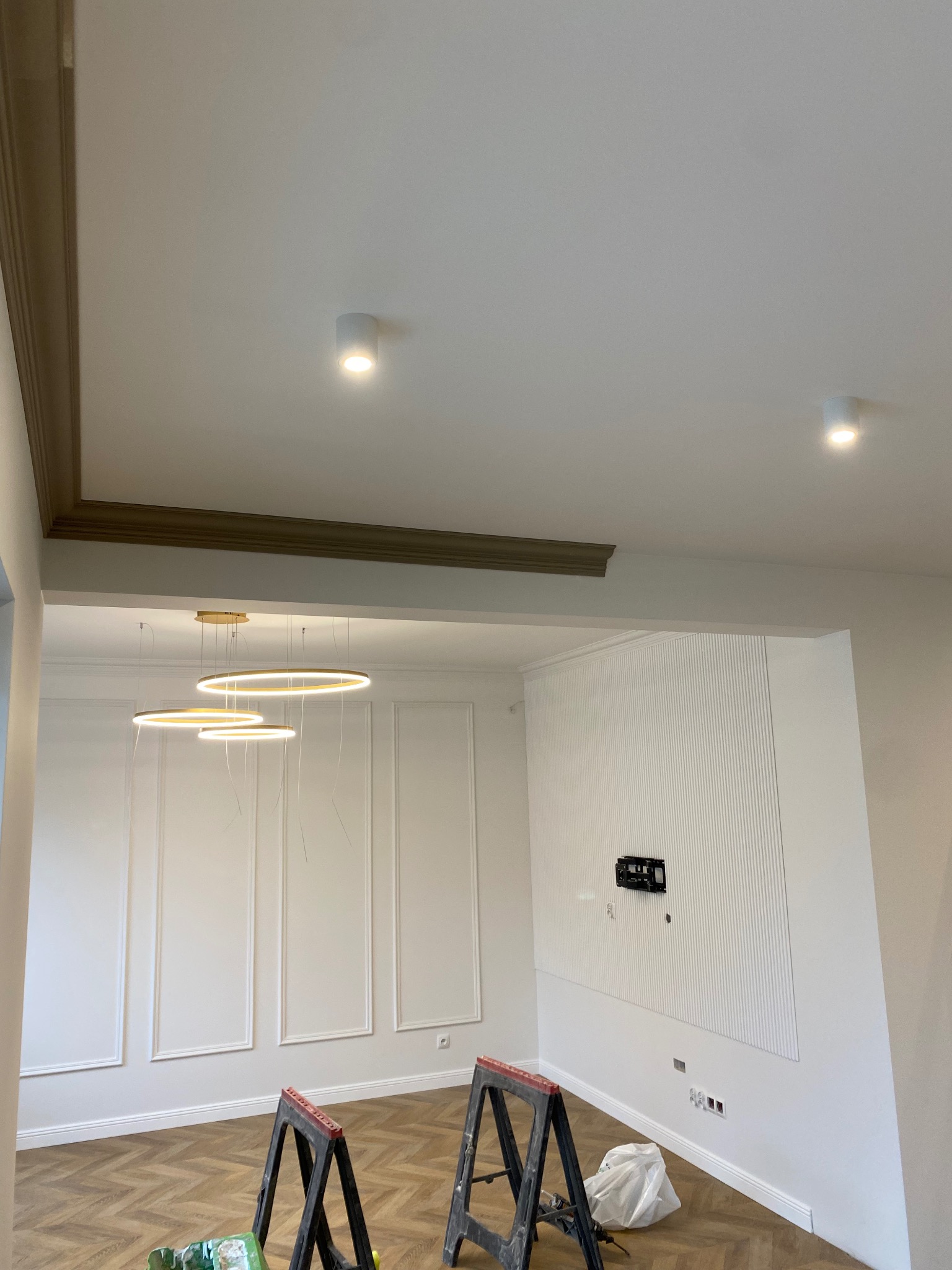 Eleganckie wnętrze w trakcie prac wykończeniowych: parkiet w jodełkę, białe ściany z dekoracyjnymi listwami, nowoczesne oświetlenie, widoczne kozły malarskie i częściowo zamontowany uchwyt...