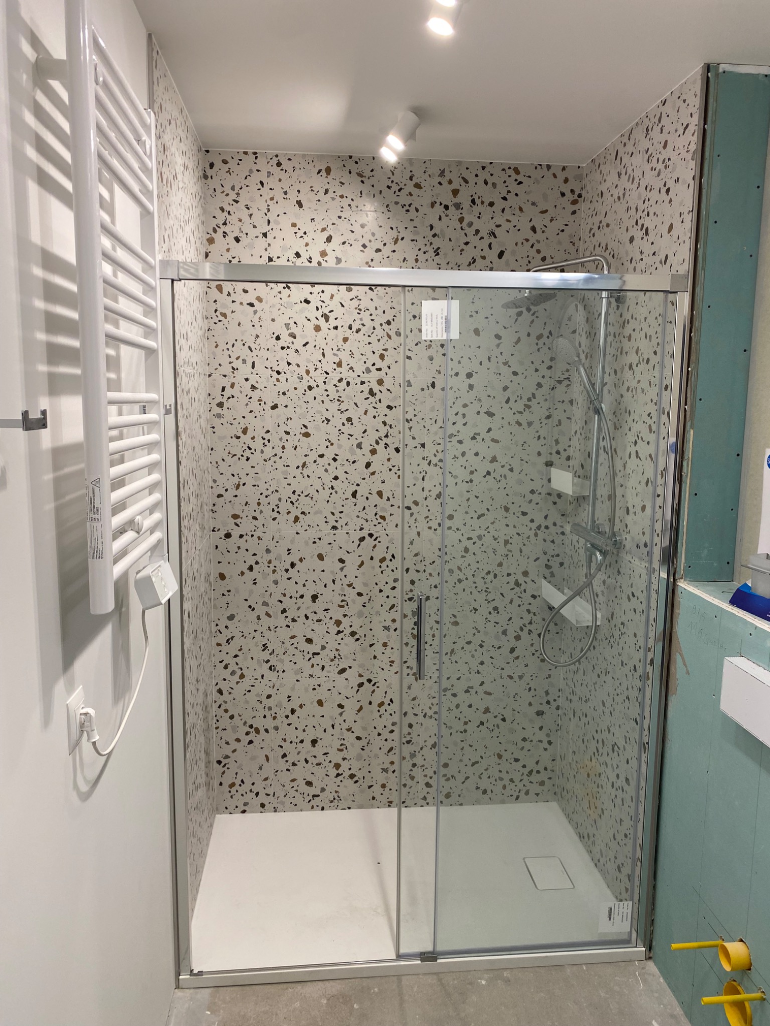 Kabina prysznicowa z przesuwnymi drzwiami, wykończona płytkami imitującymi lastryko, z grzejnikiem drabinkowym po lewej stronie i niedokończoną ścianą z płyty gipsowej po prawej.