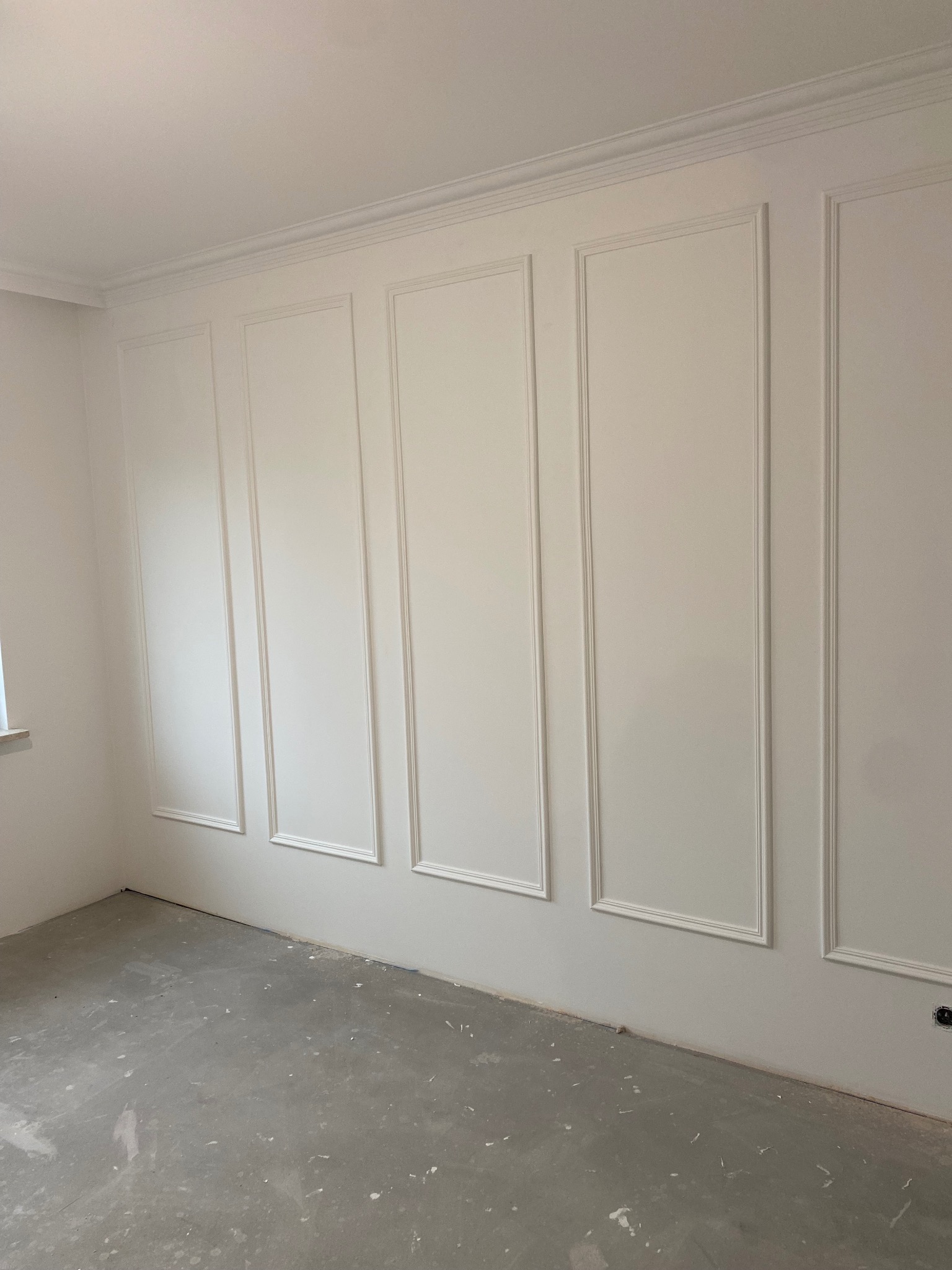 Pokój z białymi ścianami ozdobionymi sztukaterią w formie prostokątnych paneli i listew przysufitowych, surowa podłoga w odcieniach szarości, widoczne gniazdko elektryczne.