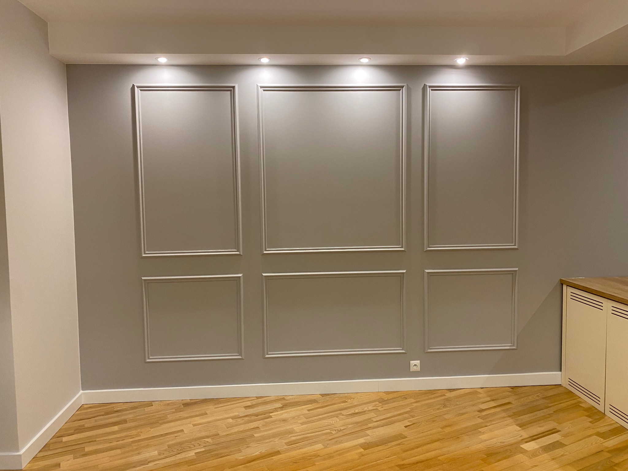 Szara ściana z dekoracyjnymi listwami w kształcie paneli, drewniana podłoga, biała listwa przypodłogowa i fragment białej szafki po prawej stronie.