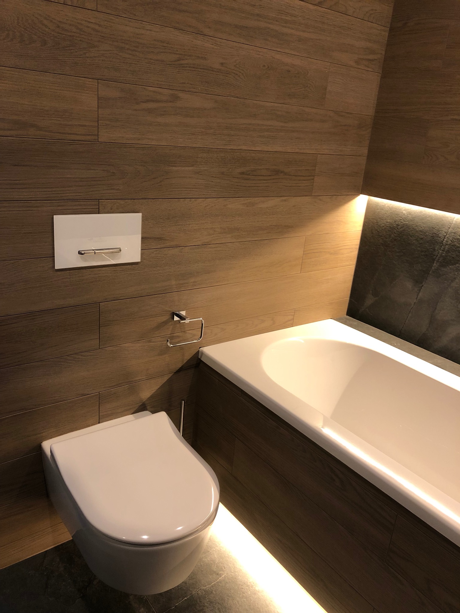 Nowoczesna łazienka z podwieszaną toaletą, wanną z oświetleniem LED i ścianami wyłożonymi płytkami imitującymi drewno.