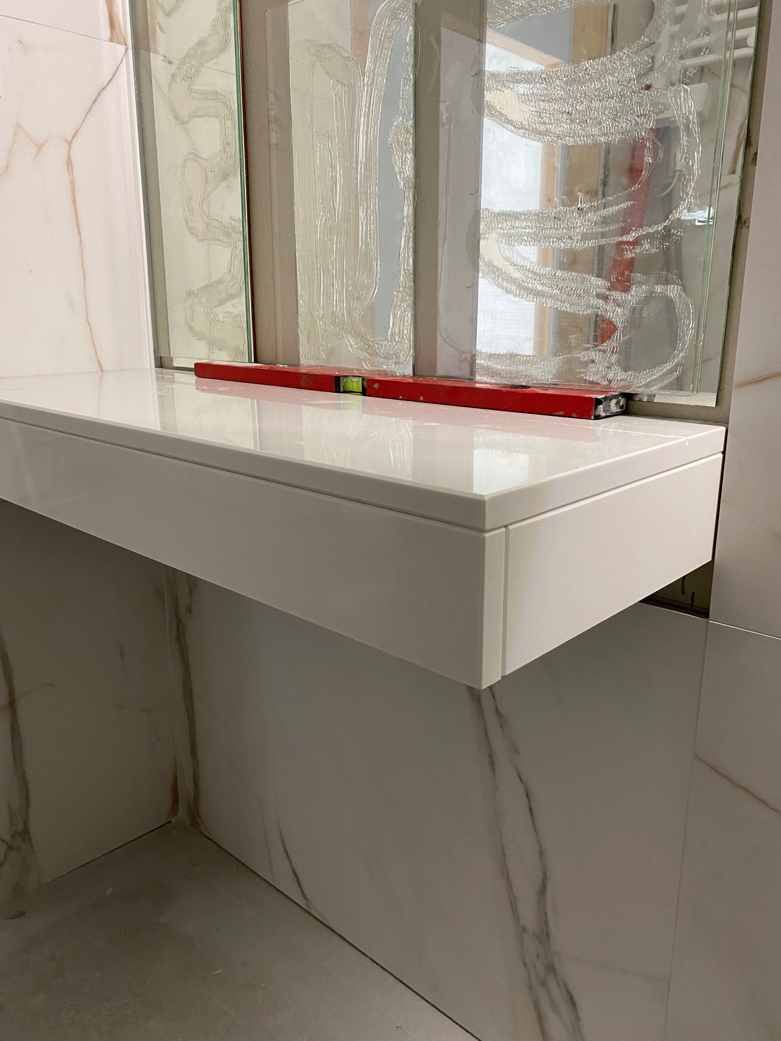 Biała, lakierowana półka pod umywalkę w łazience z marmurowymi płytkami, na której leży poziomica. Na lustrze widoczne ślady kleju po montażu.