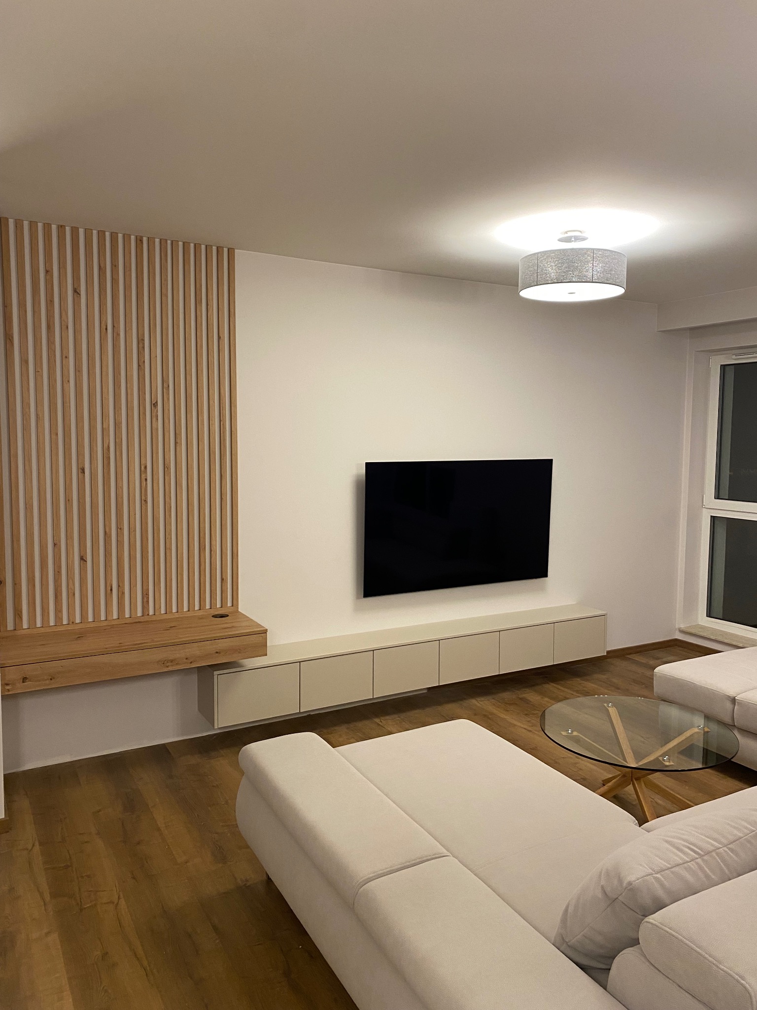 Nowoczesny salon w Warszawie z drewnianą ścianką działową, podwieszaną szafką RTV i minimalistycznym oświetleniem sufitowym nad narożną sofą.