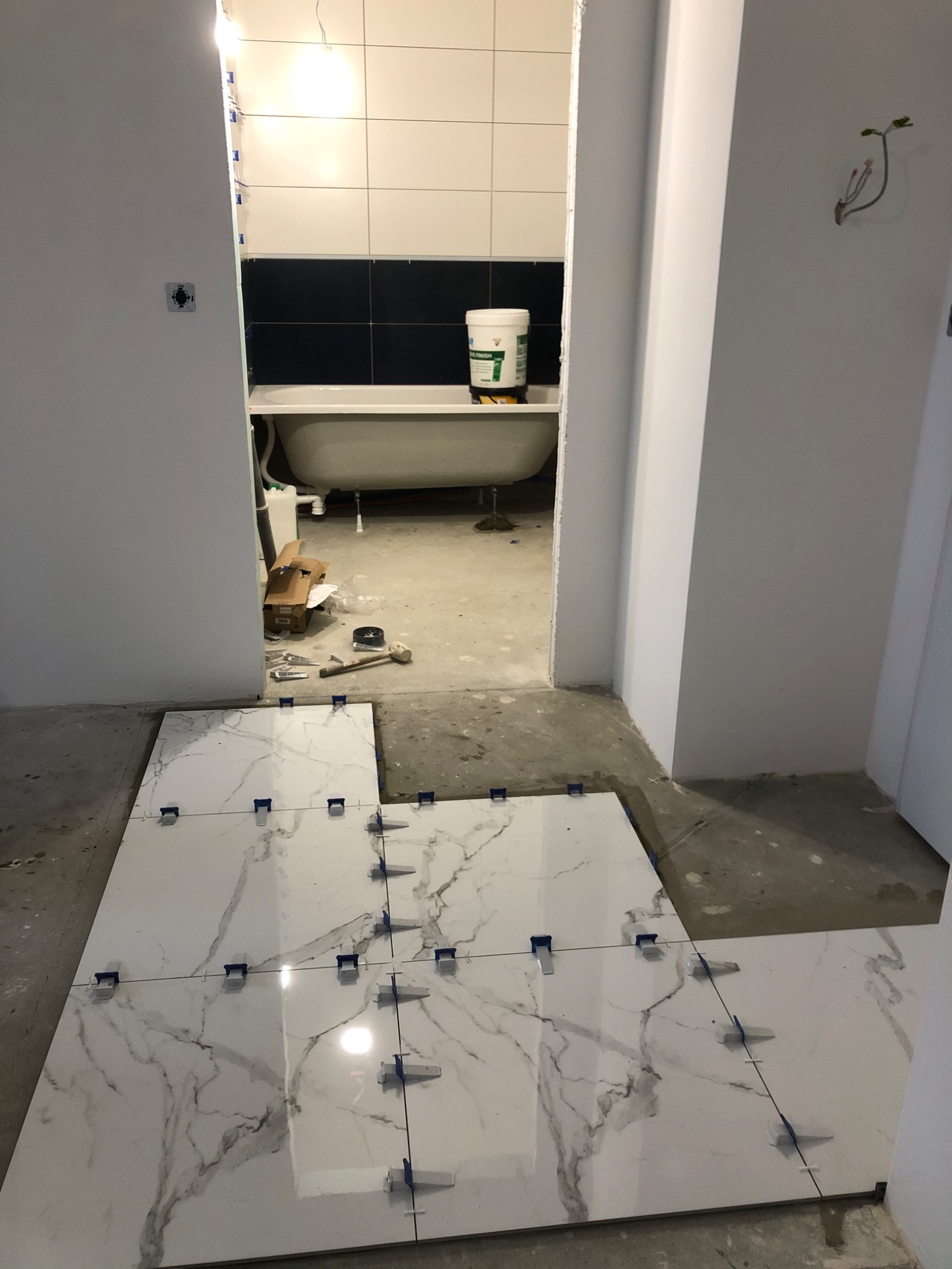 Układanie białych płytek imitujących marmur z niebieskimi krzyżykami dystansowymi na podłodze w łazience w trakcie remontu, widoczna wanna na nóżkach i biało-czarne płytki na ścianie w tle.