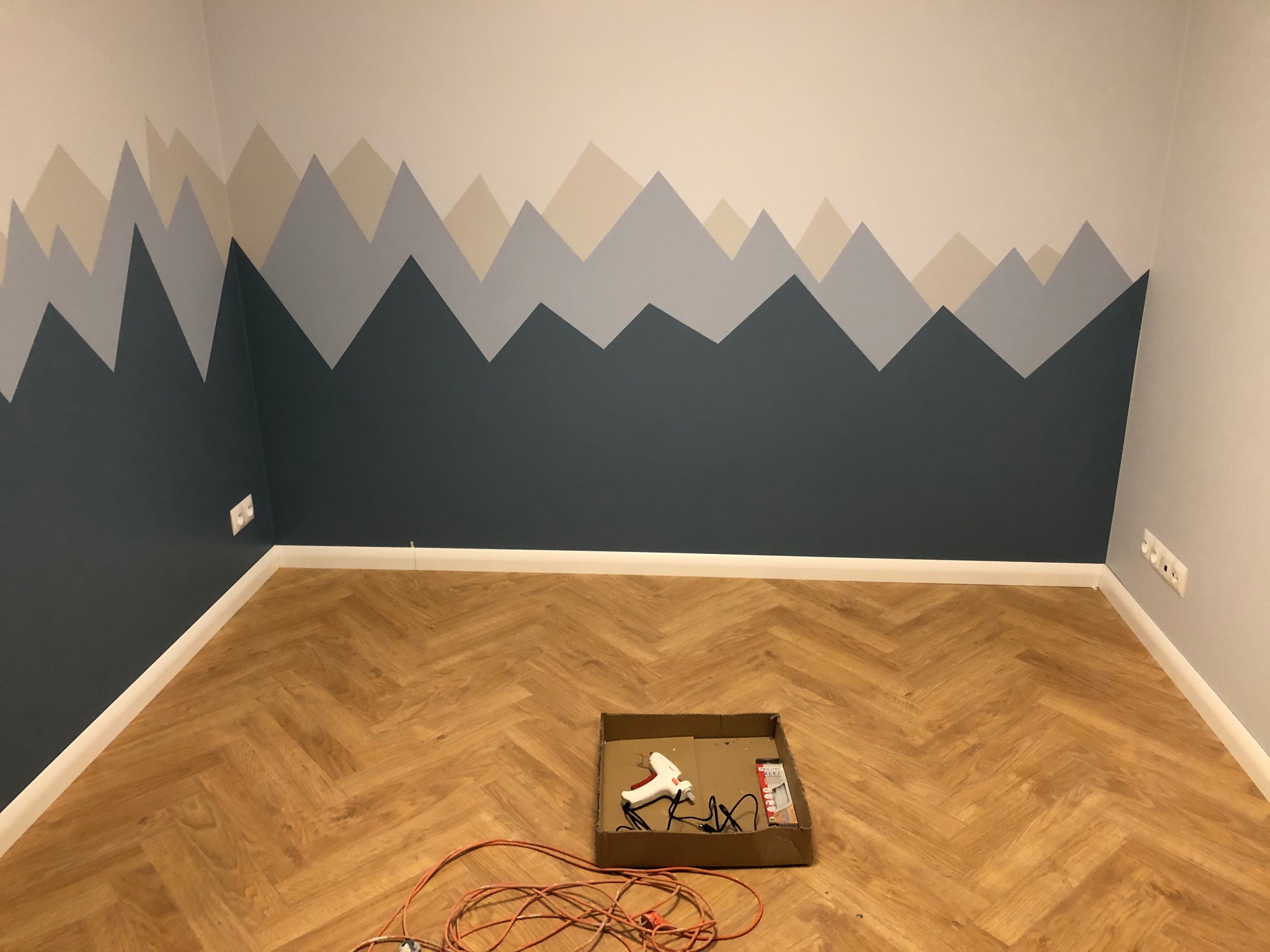 Pomieszczenie po świeżym pomalowaniu ściany w motyw górski, widoczne narzędzia malarskie w kartonie na parkiecie ułożonym w jodełkę.