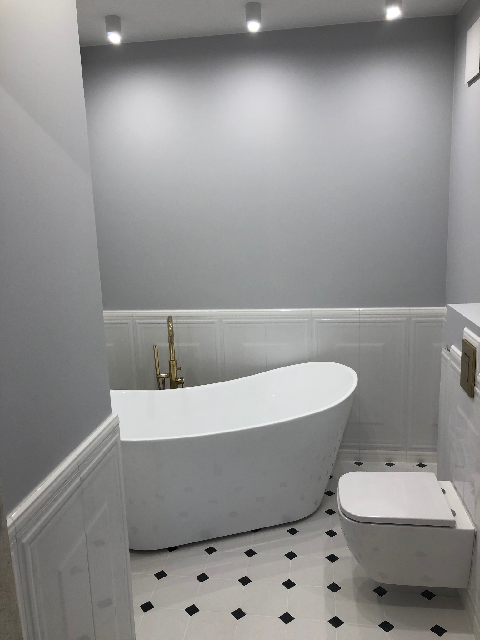 Nowoczesna łazienka w odcieniach szarości z wolnostojącą wanną z złotą armaturą i podwieszaną toaletą na podłodze w biało-czarną szachownicę.