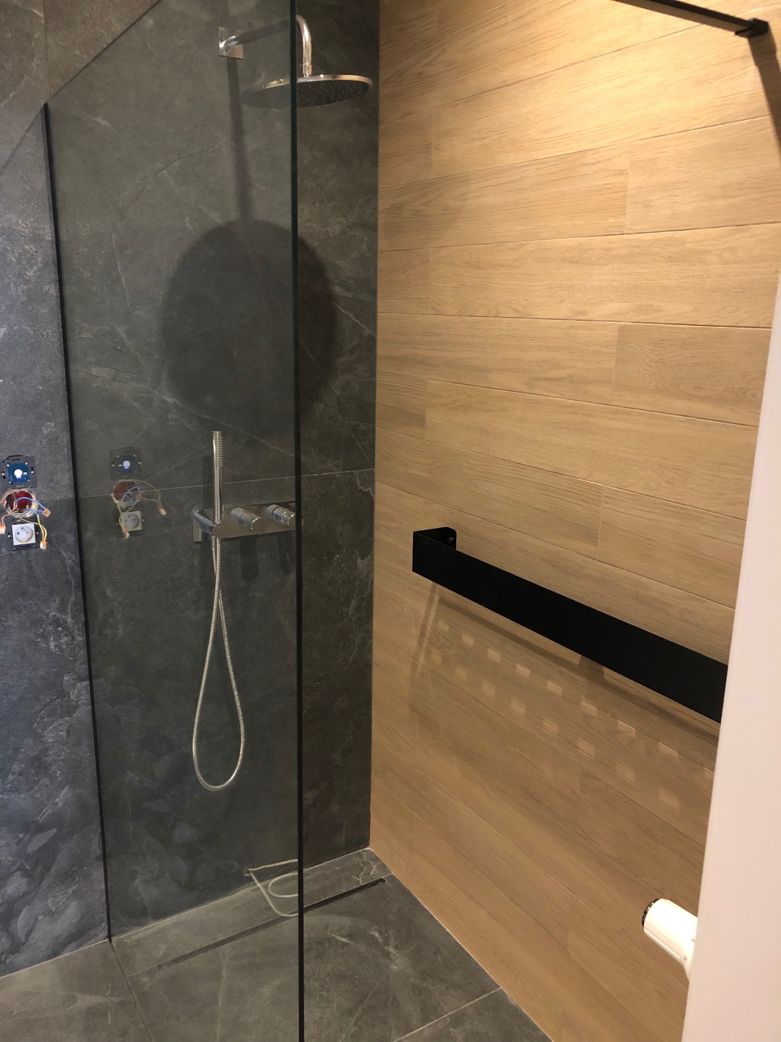 Wnętrze kabiny prysznicowej z szarą, marmurkową okładziną ścienną i podłogową, drewnopodobnymi płytkami na sąsiedniej ścianie, widoczne niezabezpieczone kable elektryczne i elementy instalacji wodnej.