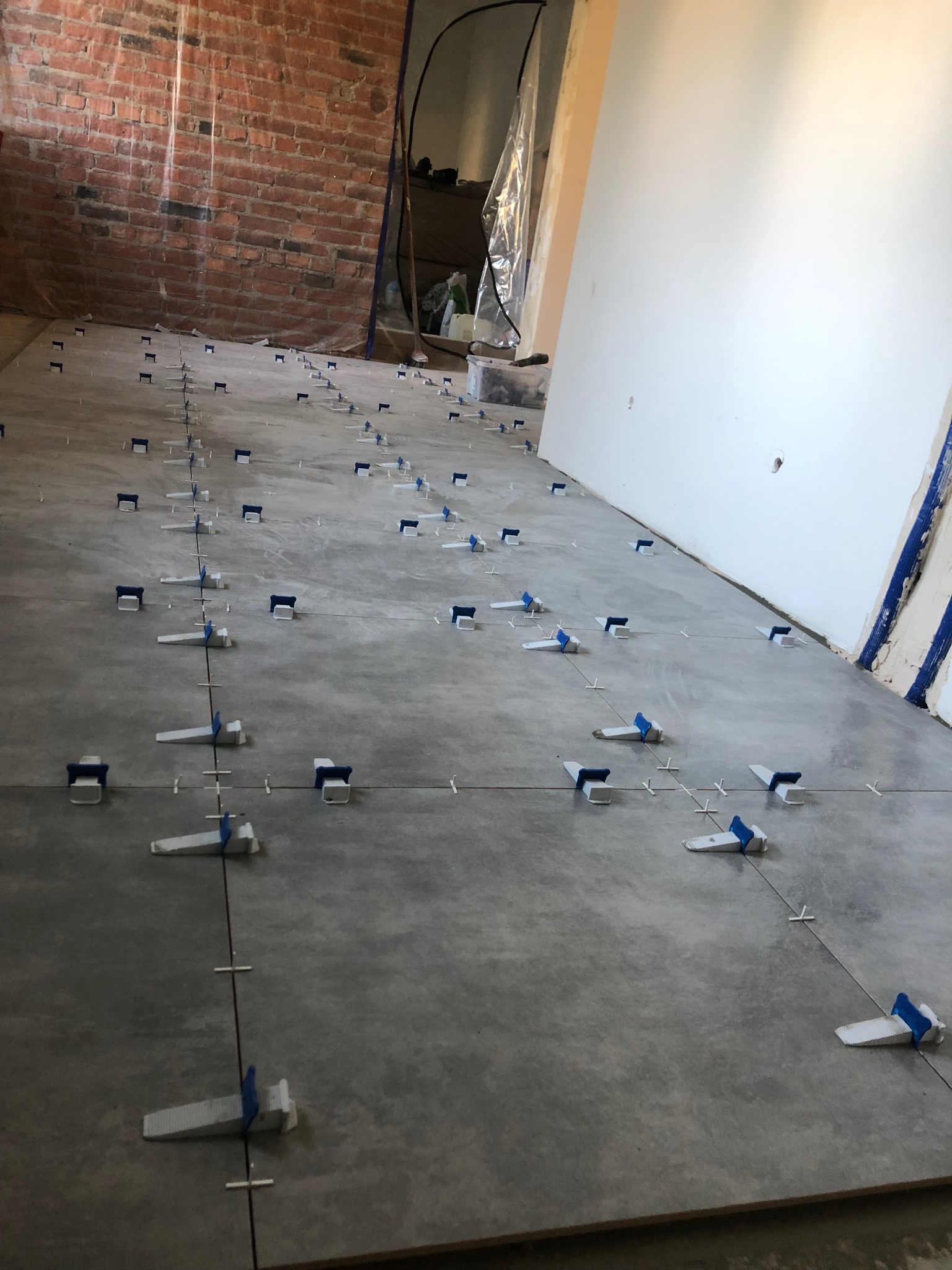 Szara podłoga w trakcie układania płytek, widoczne krzyżyki dystansowe i system poziomowania płytek z niebieskimi elementami, ściana z czerwonej cegły w tle.