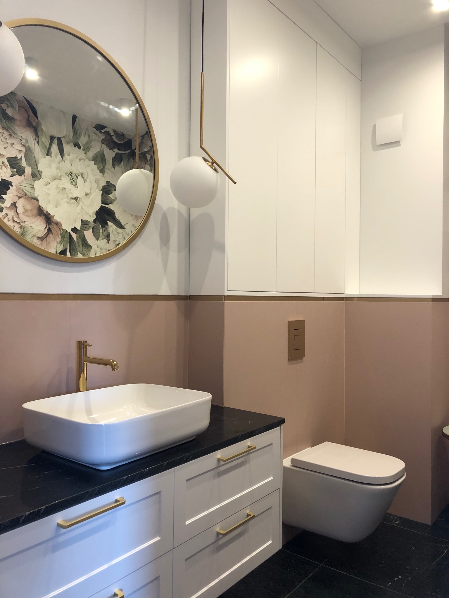Nowoczesna łazienka z umywalką nablatową na czarnym blacie, złota armatura, okrągłe lustro z kwiatowym motywem, podwieszana toaleta i różowe płytki do połowy ściany.