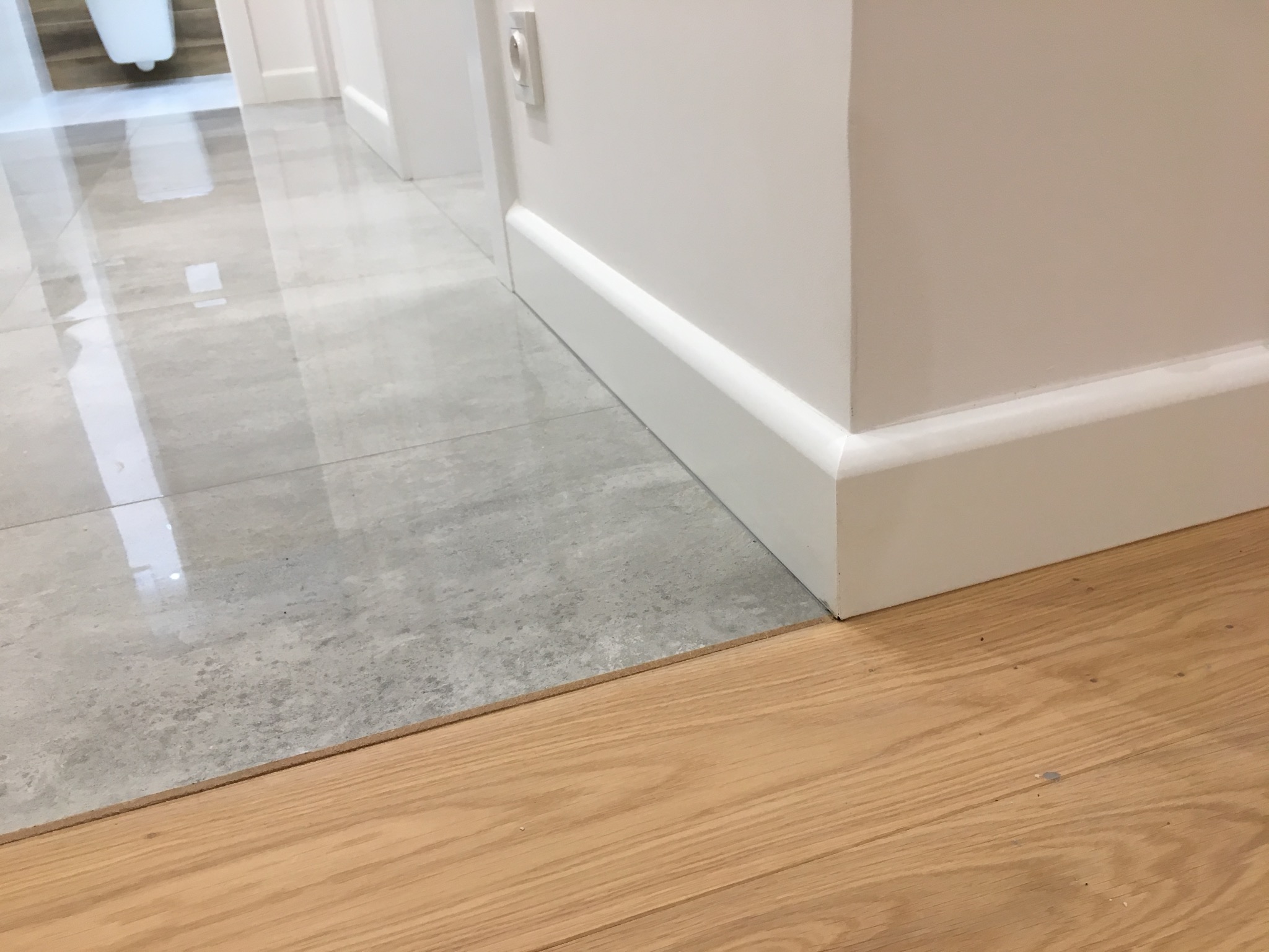 Połączenie jasnych, drewnianych paneli podłogowych z szarą, błyszczącą płytką ceramiczną w korytarzu, widoczna biała listwa przypodłogowa.