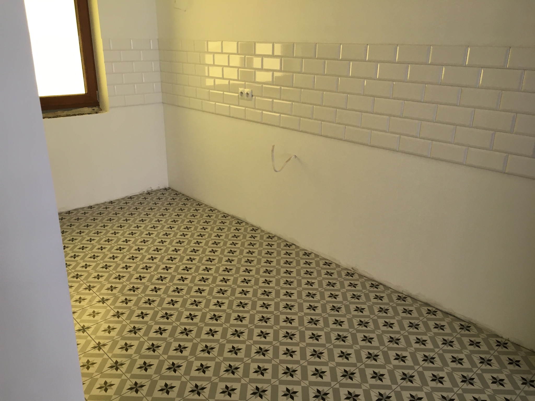 Wykończone wnętrze łazienki z nową podłogą w geometryczne wzory i białymi płytkami ściennymi typu metro, widoczne gniazdka elektryczne i wystające kable.