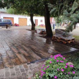 Usługi brukarskie Adam Maciąg - Ułożona nawierzchnia z kostki brukowej imitującej drewno, obrzeża z czarnego tworzywa, obok rabata z różowymi kwiatami, w tle zaparkowany samochód i budynek gospodarczy.