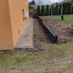 Usługi brukarskie Adam Maciąg - Ceglana ścieżka prowadząca wzdłuż pomarańczowego budynku do przygotowanego terenu z czarnym obrzeżem, w tle rząd tuj i szare budynki gospodarcze.