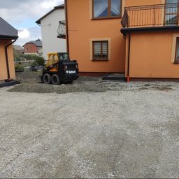 Usługi brukarskie Adam Maciąg - Przygotowanie podłoża pod kostkę brukową: ubijarka wibracyjna, miniładowarka Bobcat i zagęszczarka płytowa na żwirowym podłożu przed budynkami o pomarańczowej elewacji.