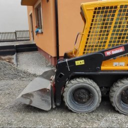 Usługi brukarskie Adam Maciąg - Żółta miniładowarka Bobcat S100 na placu budowy, przygotowującym teren pod kostkę brukową. Widoczne obrzeża i podsypka z kruszywa.
