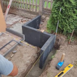 Usługi brukarskie Adam Maciąg - Wykop pod fundament z ustawionymi szalunkami z czarnych płyt, wytyczony sznurkiem, obok leżą poziomica i gumowy młotek, w tle widoczny częściowo ułożony bruk i ogrodzenie.
