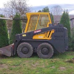 Usługi brukarskie Adam Maciąg - Żółto-czarna miniładowarka Bobcat S100 na trawniku, obok pracownik z łopatą i fragmenty kostki brukowej, widoczne tuje i domy w tle.