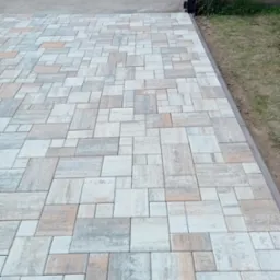 Ułożona nawierzchnia z kostki brukowej w odcieniach szarości, beżu i bieli, z widocznym obrzeżem i fragmentem trawnika oraz ogrodzenia w tle.