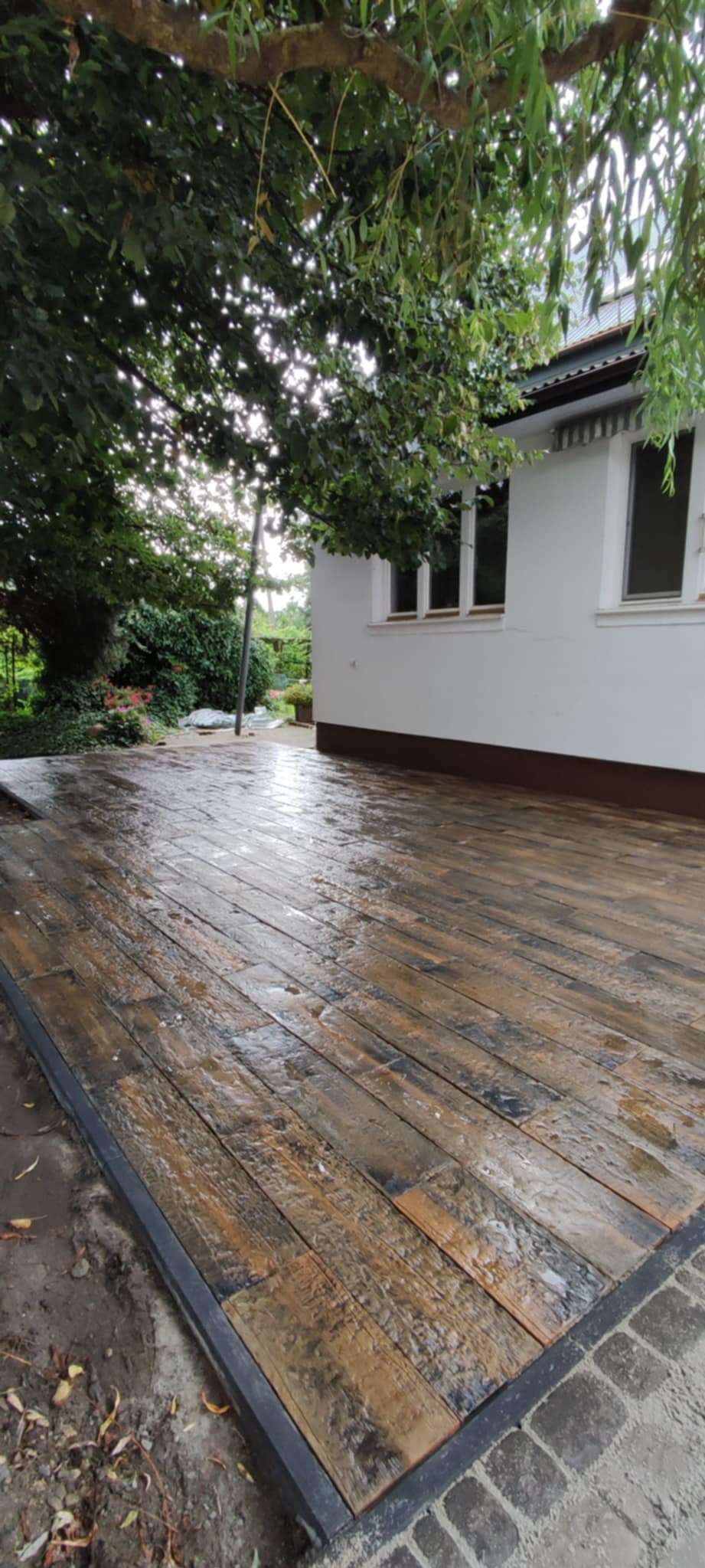 Ułożona, mokra nawierzchnia z kostki brukowej imitującej drewno, obramowana metalową listwą i kostką brukową, przylegająca do białego budynku z ogrodem w tle.