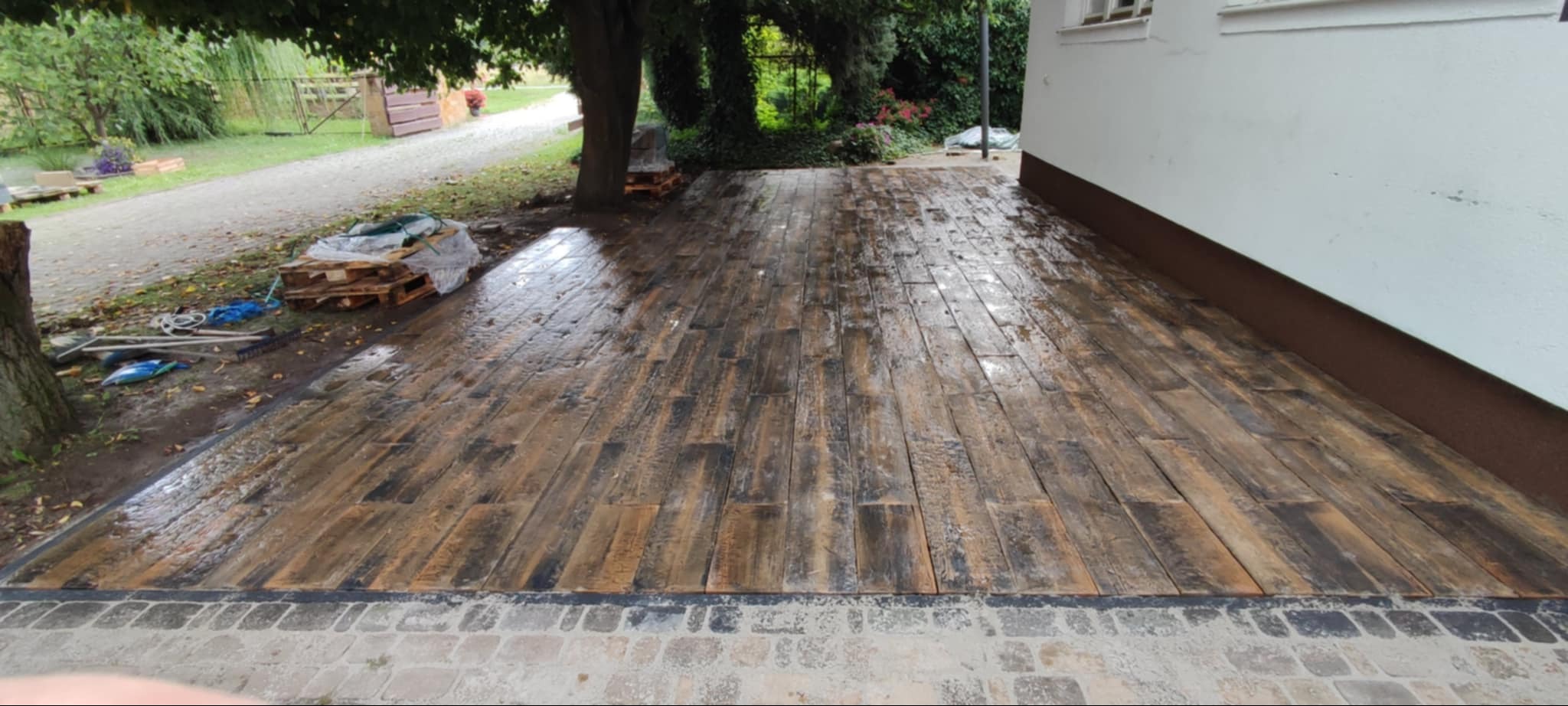 Wykończony podjazd z kostki brukowej imitującej drewno, obrzeże z kostki granitowej, widoczne palety i narzędzia w tle.