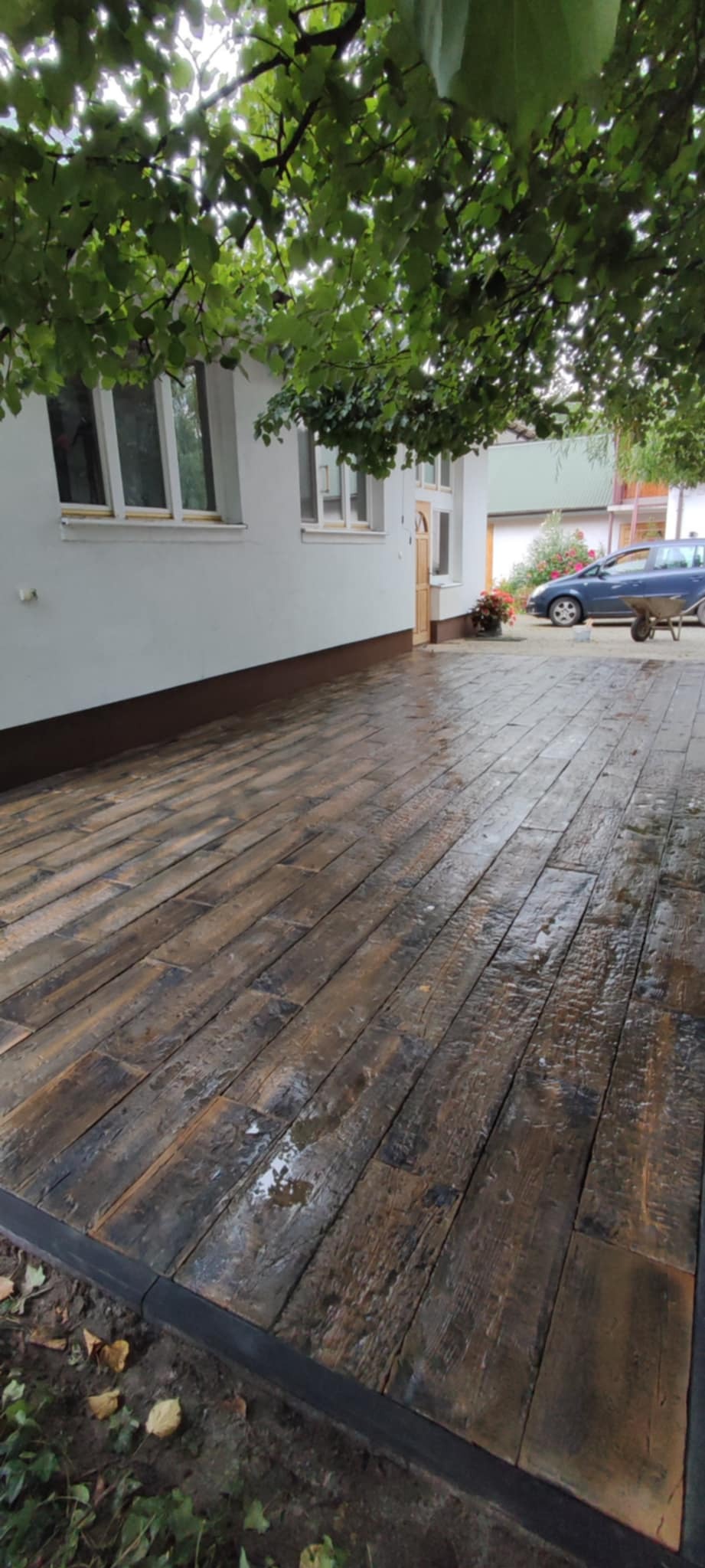 Świeżo ułożona, mokra kostka brukowa imitująca drewniane deski, obramowana czarnym krawężnikiem, na tle białego domu z brązową podmurówką i zieleni drzew.