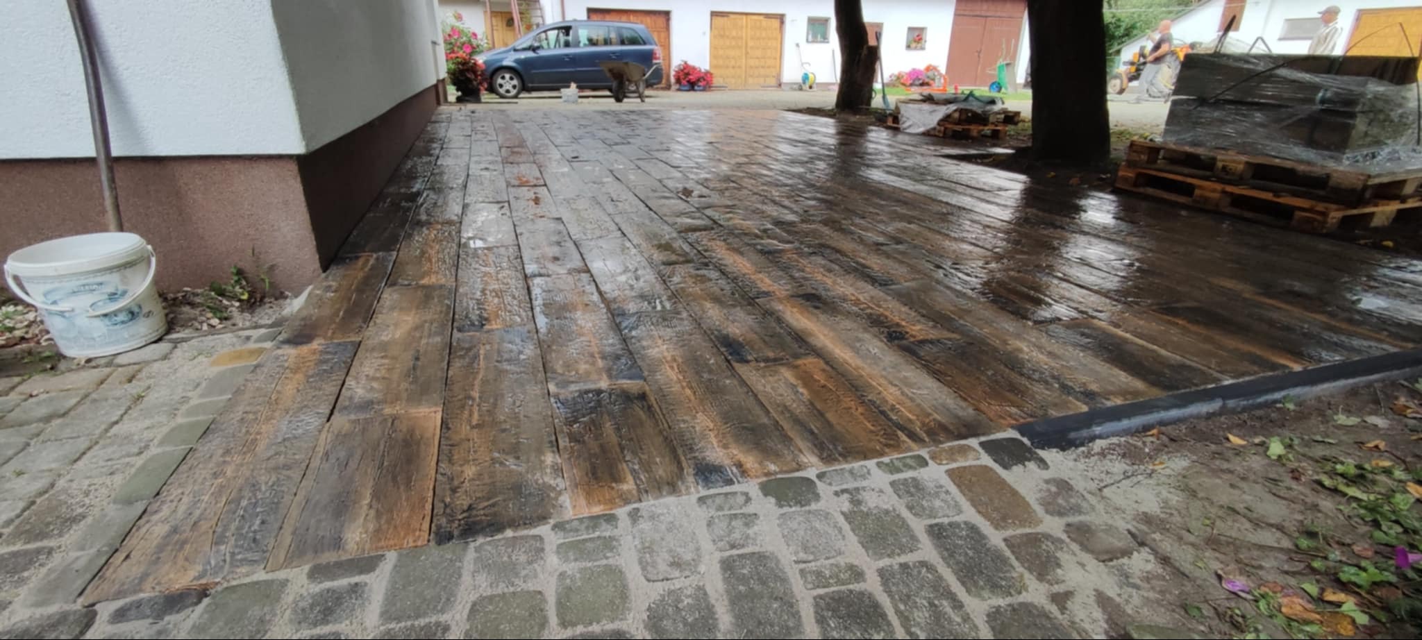Ułożona nawierzchnia z desek kompozytowych imitujących drewno, obrzeża z kostki granitowej, widoczne palety z materiałem budowlanym w tle.