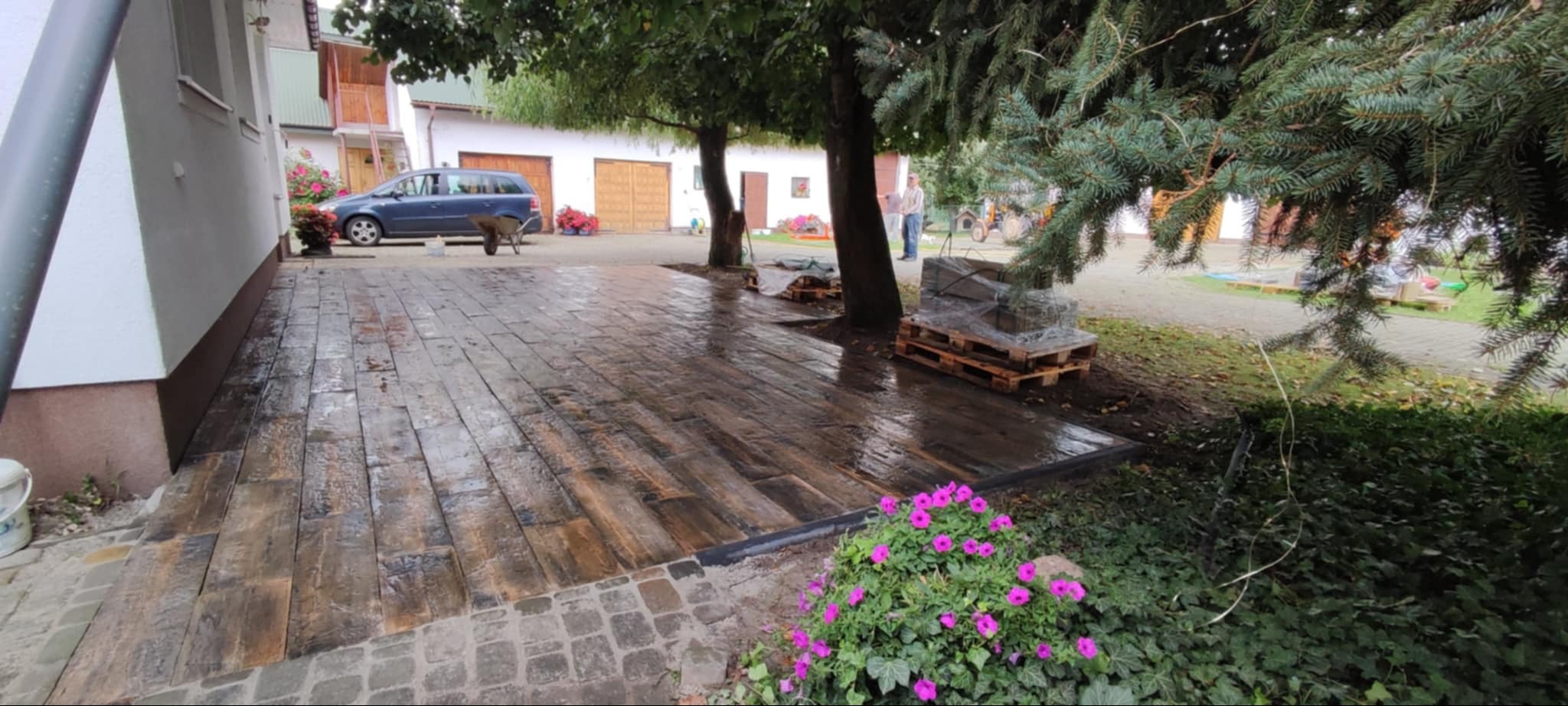 Ułożona nawierzchnia z kostki brukowej imitującej drewno, obrzeża z czarnego tworzywa, obok rabata z różowymi kwiatami, w tle zaparkowany samochód i budynek gospodarczy.