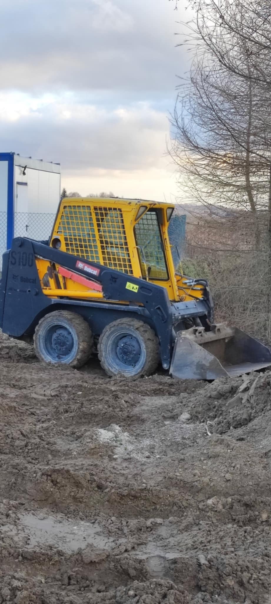 Żółto-czarna ładowarka Bobcat S100 na placu budowy, z widocznymi śladami błota na oponach i łyżce, w tle ogrodzenie z siatki i drzewa bez liści.