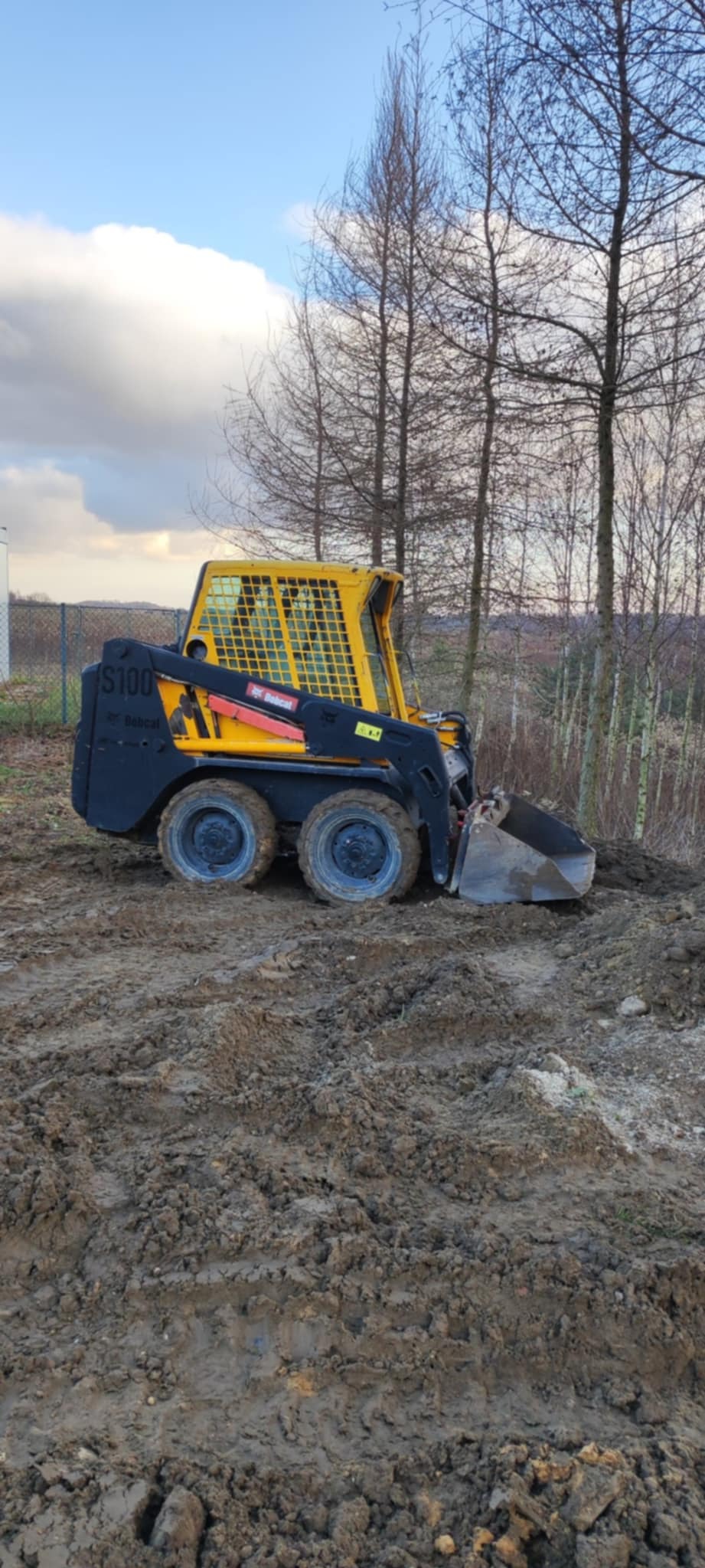 Żółto-czarna miniładowarka Bobcat S100 na placu budowy z ubitym błotem, w tle drzewa bez liści i fragment ogrodzenia.