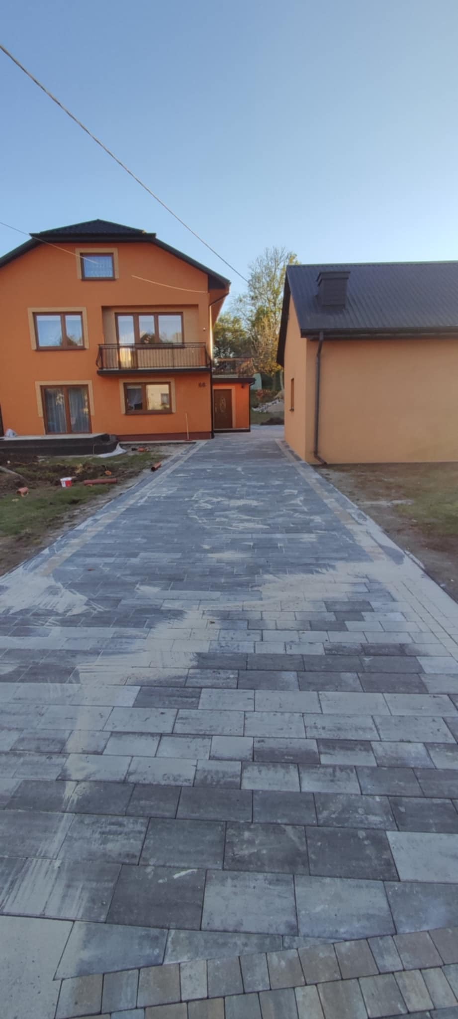 Ułożona szara kostka brukowa prowadząca do pomarańczowego domu z balkonem, widoczne ślady po piasku i resztki ziemi po bokach.