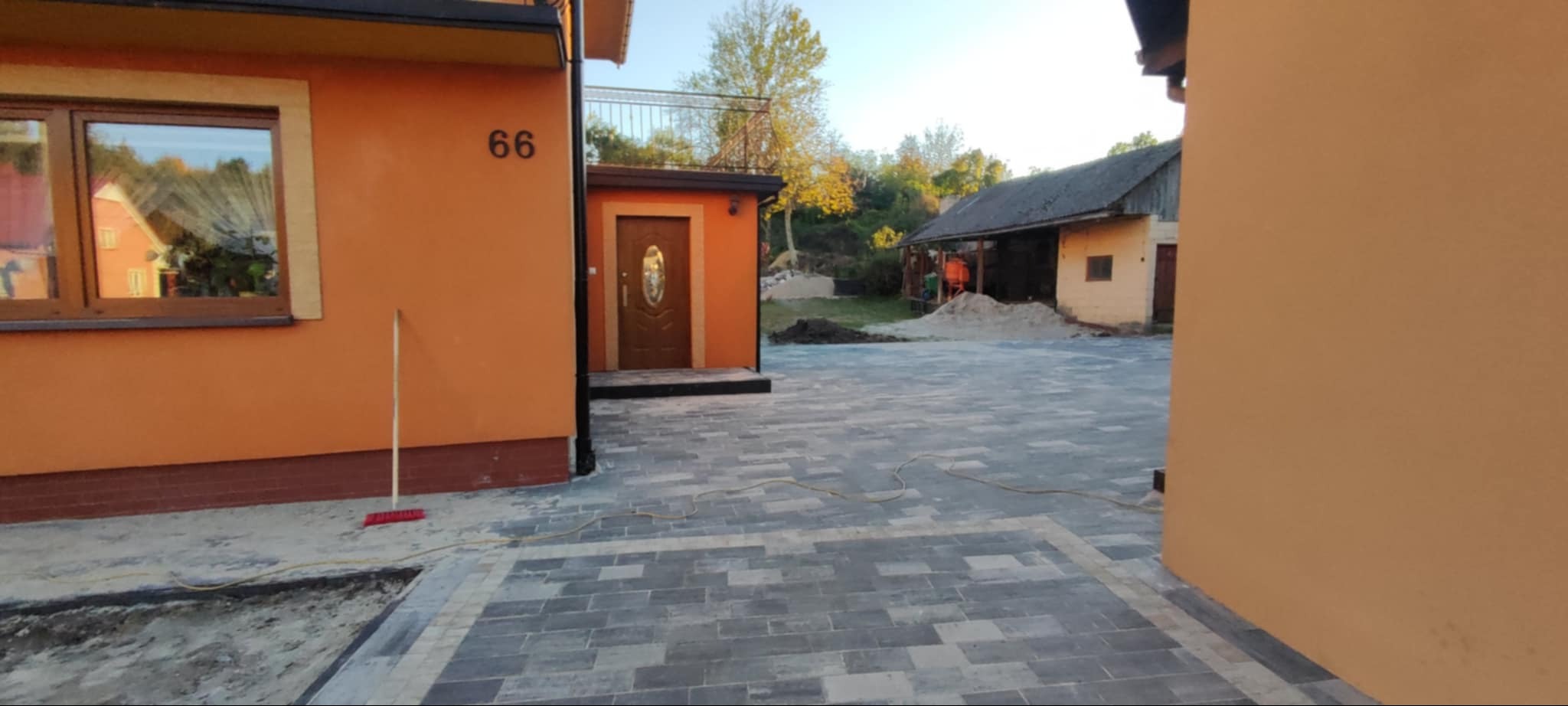 Ułożona szara kostka brukowa na podwórku domu z pomarańczową elewacją i numerem 66, widoczny budynek gospodarczy w tle i rozłożona warstwa piasku.