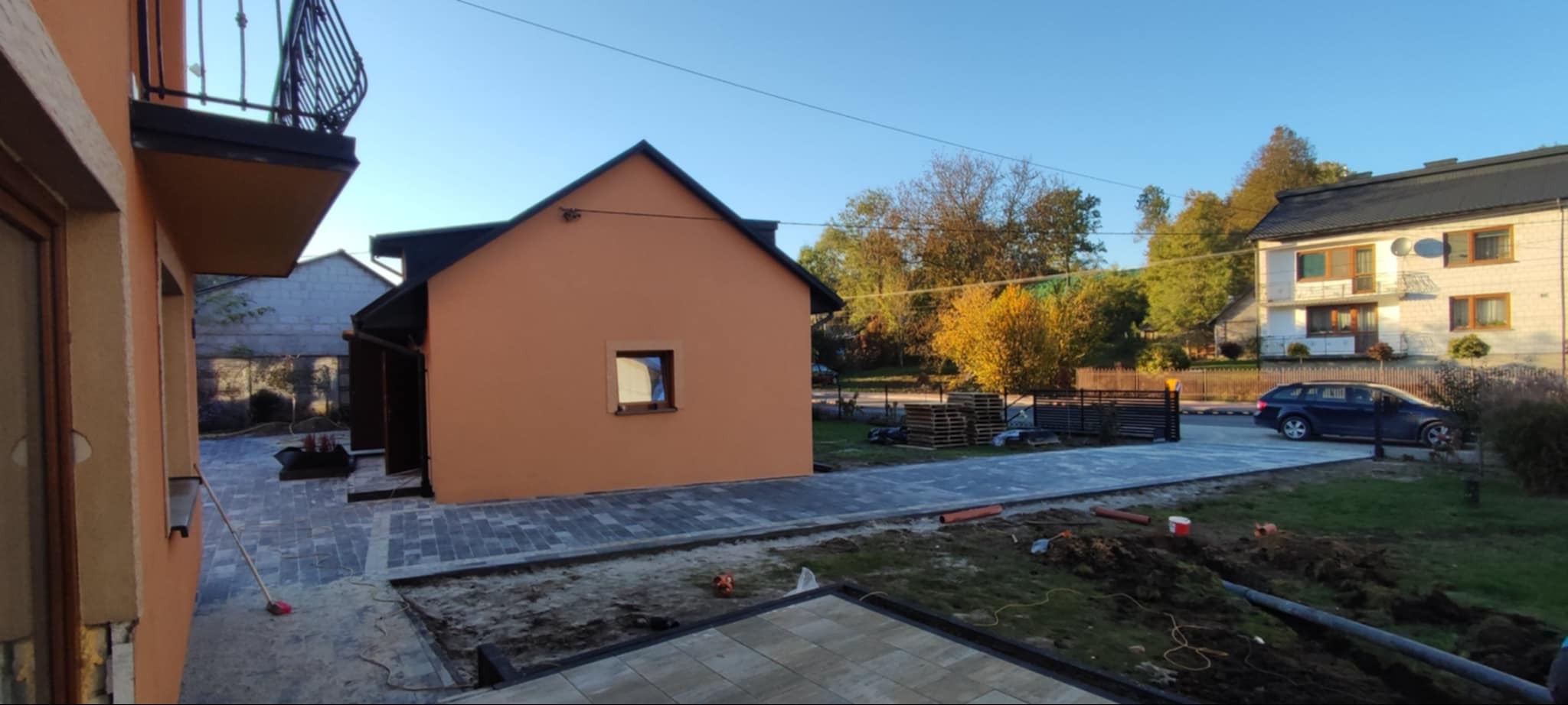 Ułożona kostka brukowa w kolorze szarym na posesji z pomarańczowym budynkiem gospodarczym, widoczne palety, częściowo wykopana ziemia i samochód osobowy w tle.