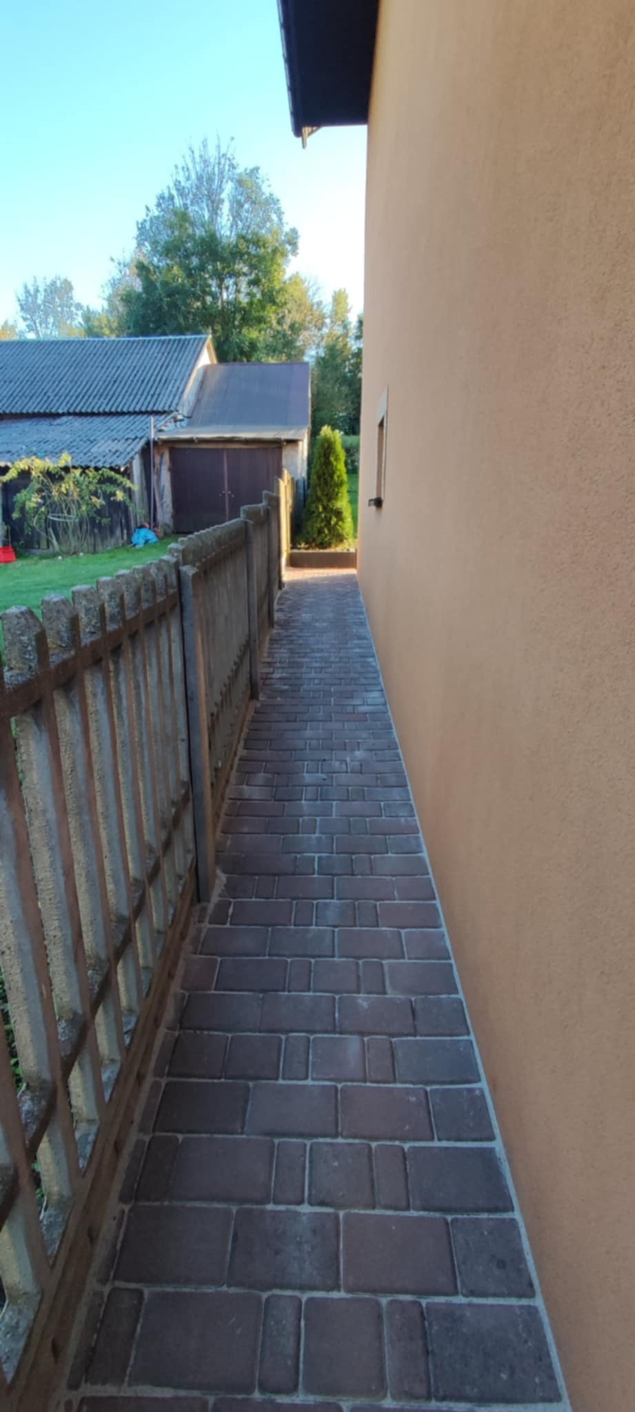 Ułożona ścieżka z kostki brukowej prowadząca wzdłuż ściany budynku, widoczny fragment ogrodzenia i zieleń w tle.