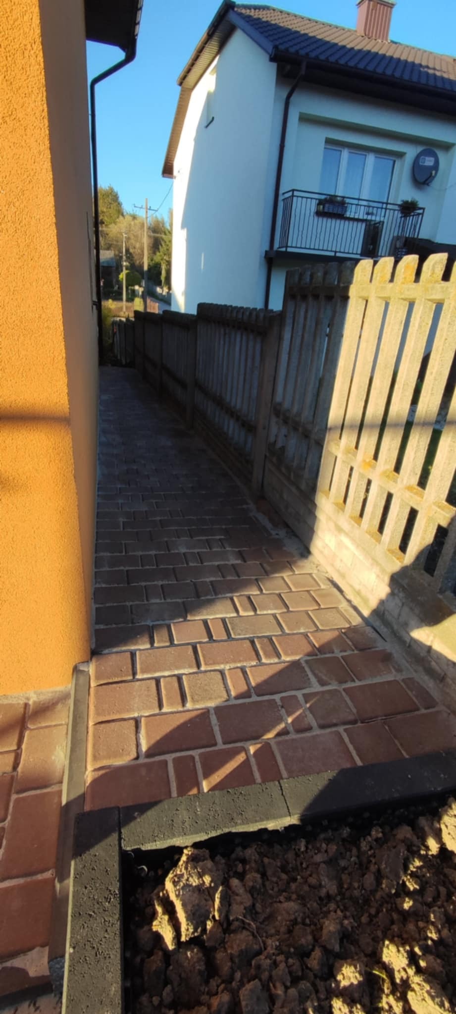 Ceglana ścieżka z kostki prowadząca wzdłuż ogrodzenia i budynku, z widocznym fragmentem wykopu na obrzeżu.