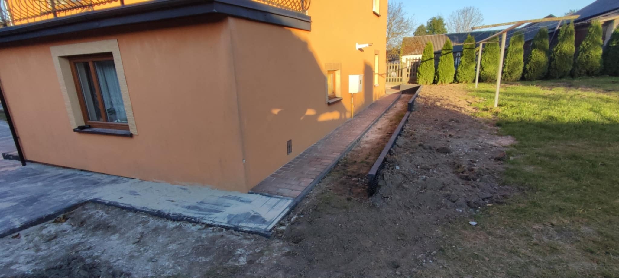 Ułożona ścieżka z czerwonej kostki prowadząca wzdłuż domu w Klimontowie, z widocznym fragmentem tarasu z szarej kostki i przygotowanym terenem pod dalsze prace ziemne.