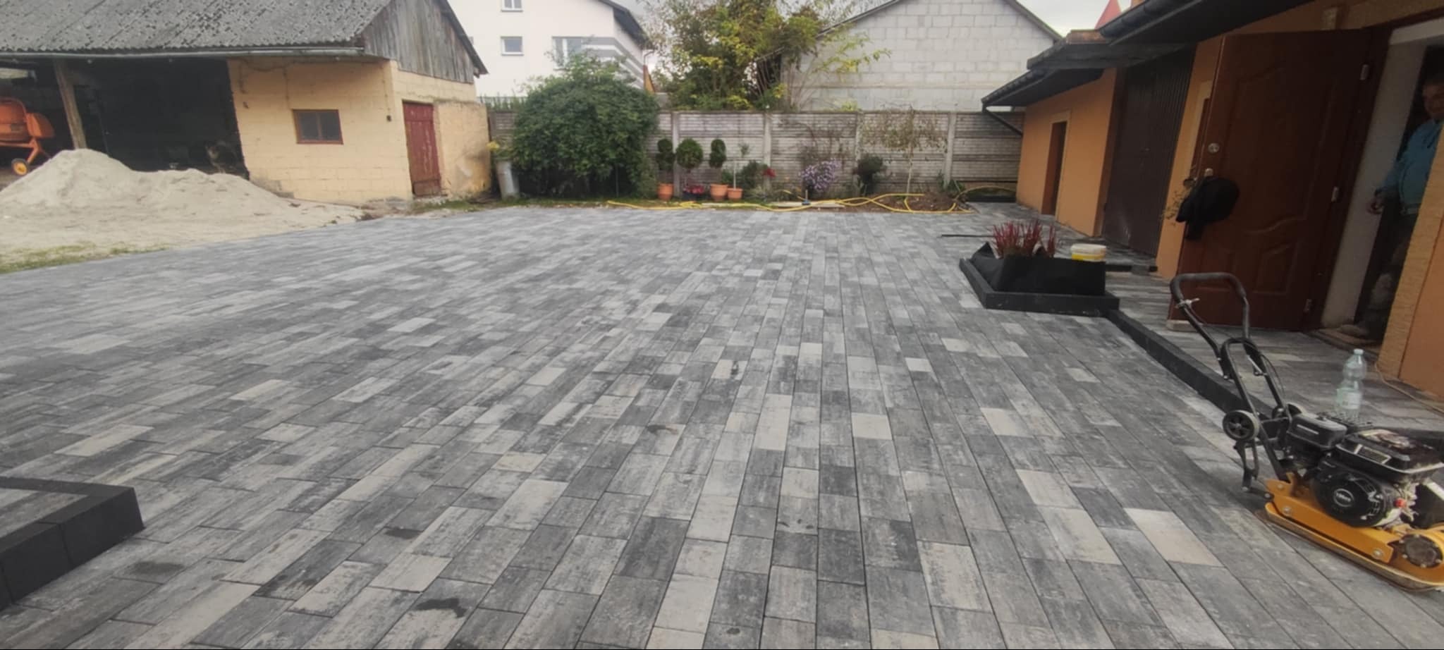 Ułożona szara kostka brukowa w jodełkę na posesji, z widoczną ubijarką wibracyjną, stertą piasku i betoniarką w tle.
