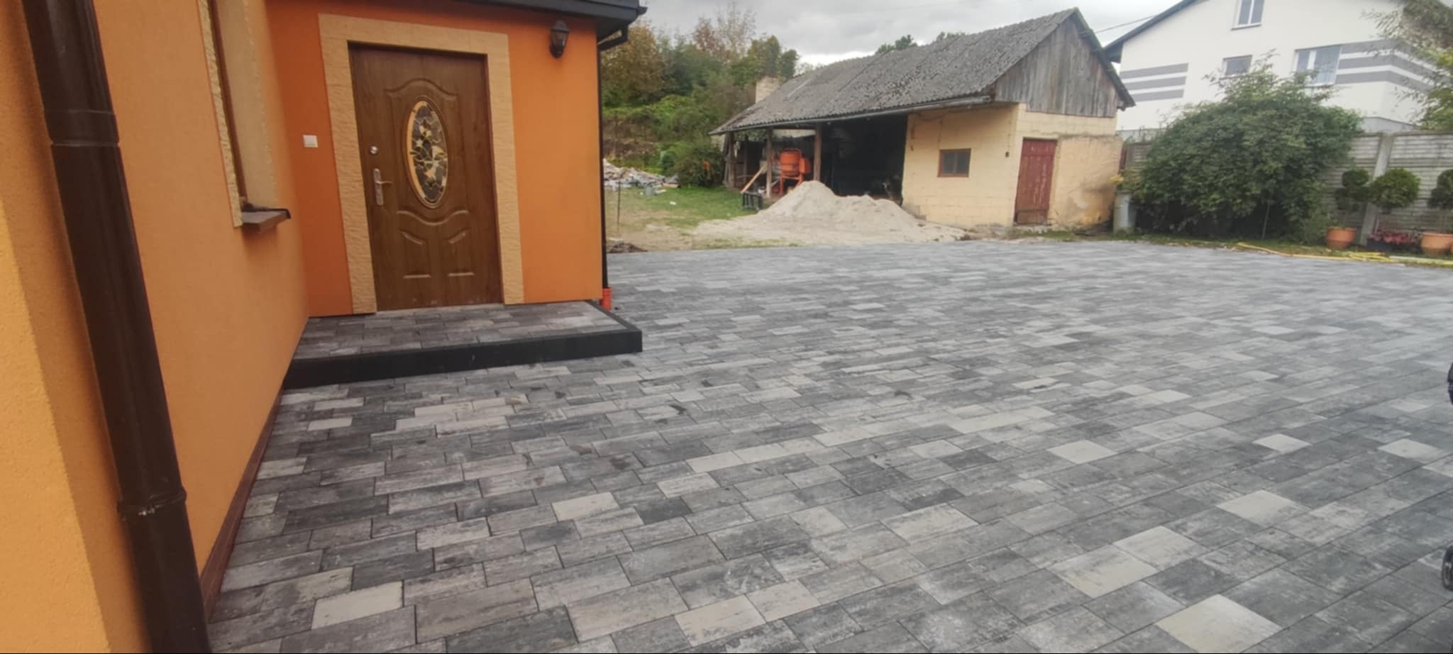 Ułożona szara kostka brukowa przed pomarańczowym domem z drewnianymi drzwiami; w tle widoczny stary budynek gospodarczy z hałdą piasku.