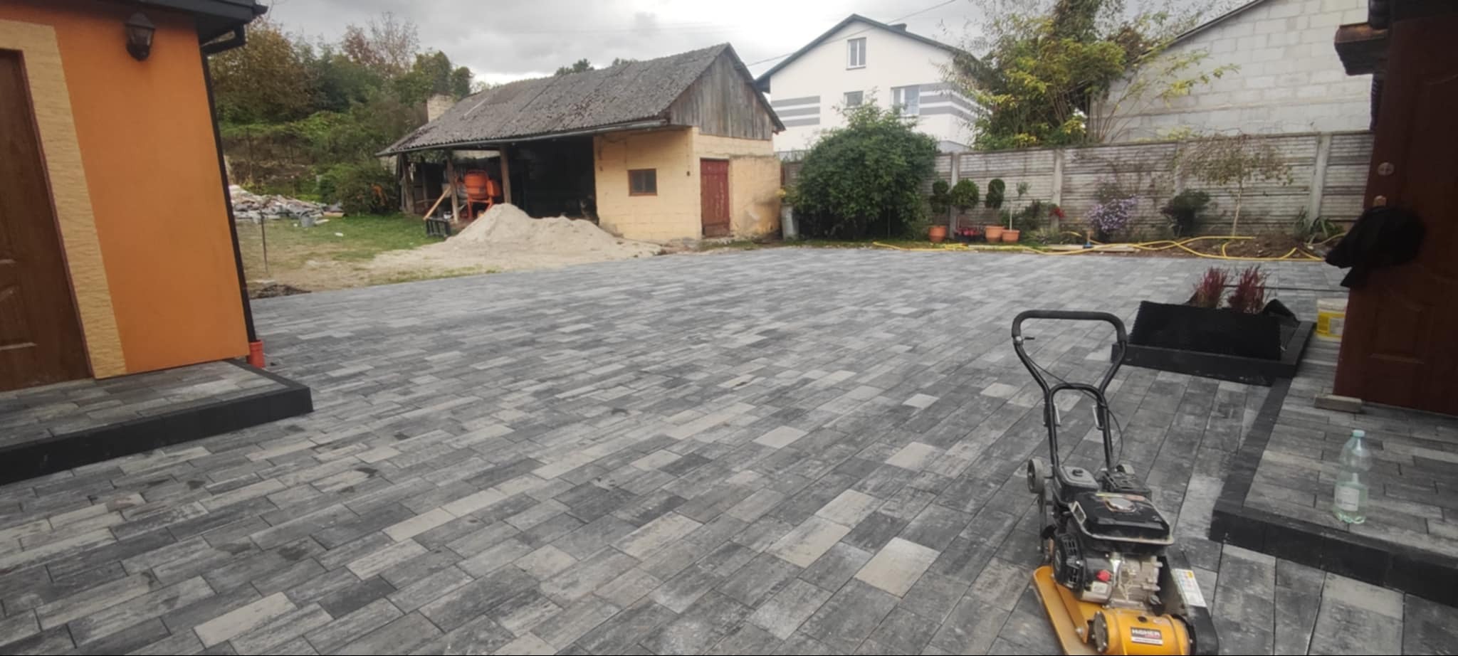 Ułożona szara kostka brukowa w jodełkę na posesji, widoczna zagęszczarka w prawym dolnym rogu, w tle budynek gospodarczy z mieszalnikiem betonu i stertą piasku.
