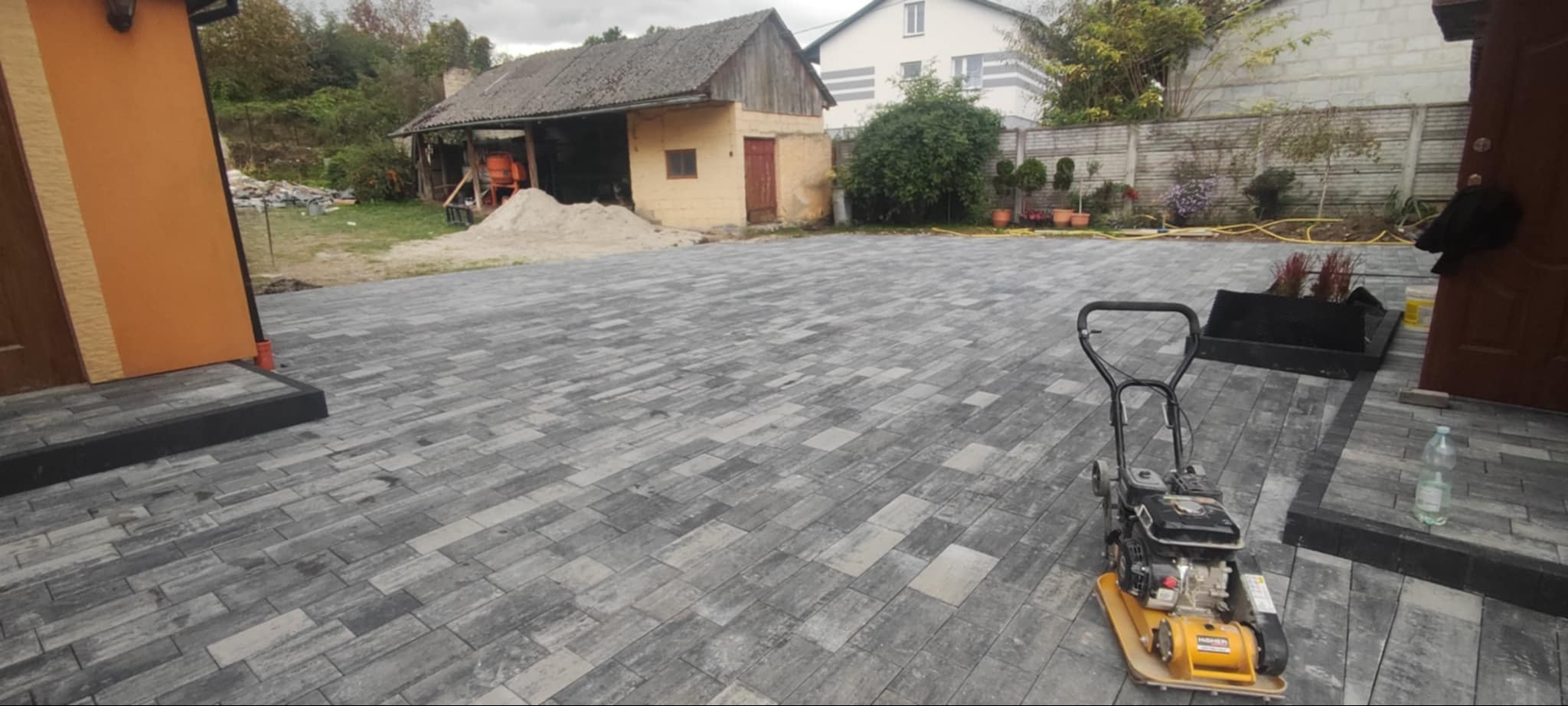 Ukończony podjazd z szarej kostki brukowej ułożonej w jodełkę, widoczna zagęszczarka spalinowa, w tle budynek gospodarczy i dom jednorodzinny.