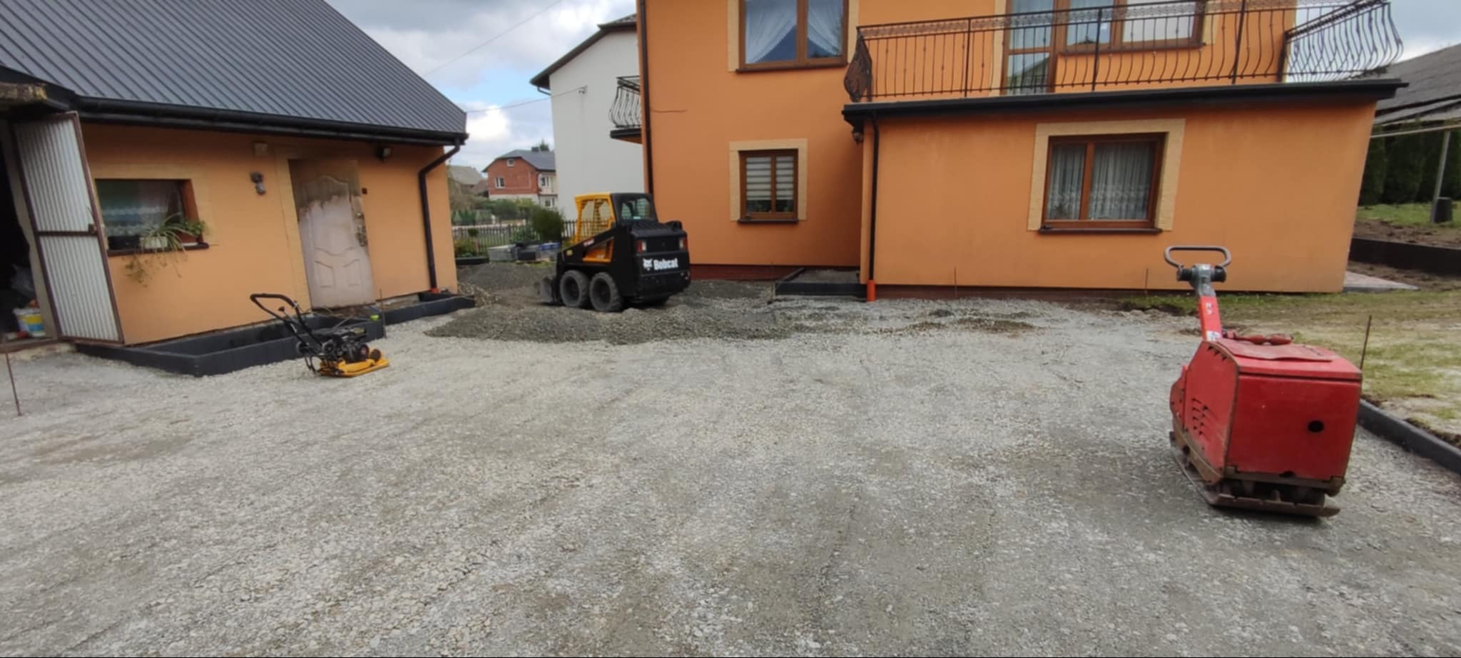 Przygotowanie podłoża pod kostkę brukową: ubijarka wibracyjna, miniładowarka Bobcat i zagęszczarka płytowa na żwirowym podłożu przed budynkami o pomarańczowej elewacji.