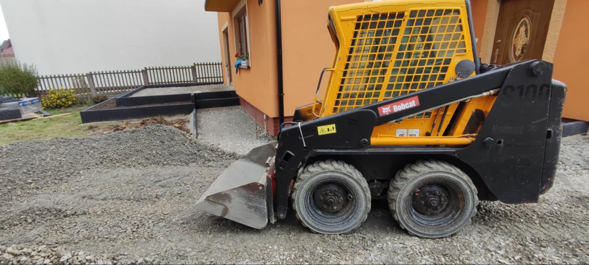 Żółta miniładowarka Bobcat S100 na placu budowy, przygotowującym teren pod kostkę brukową. Widoczne obrzeża i podsypka z kruszywa.