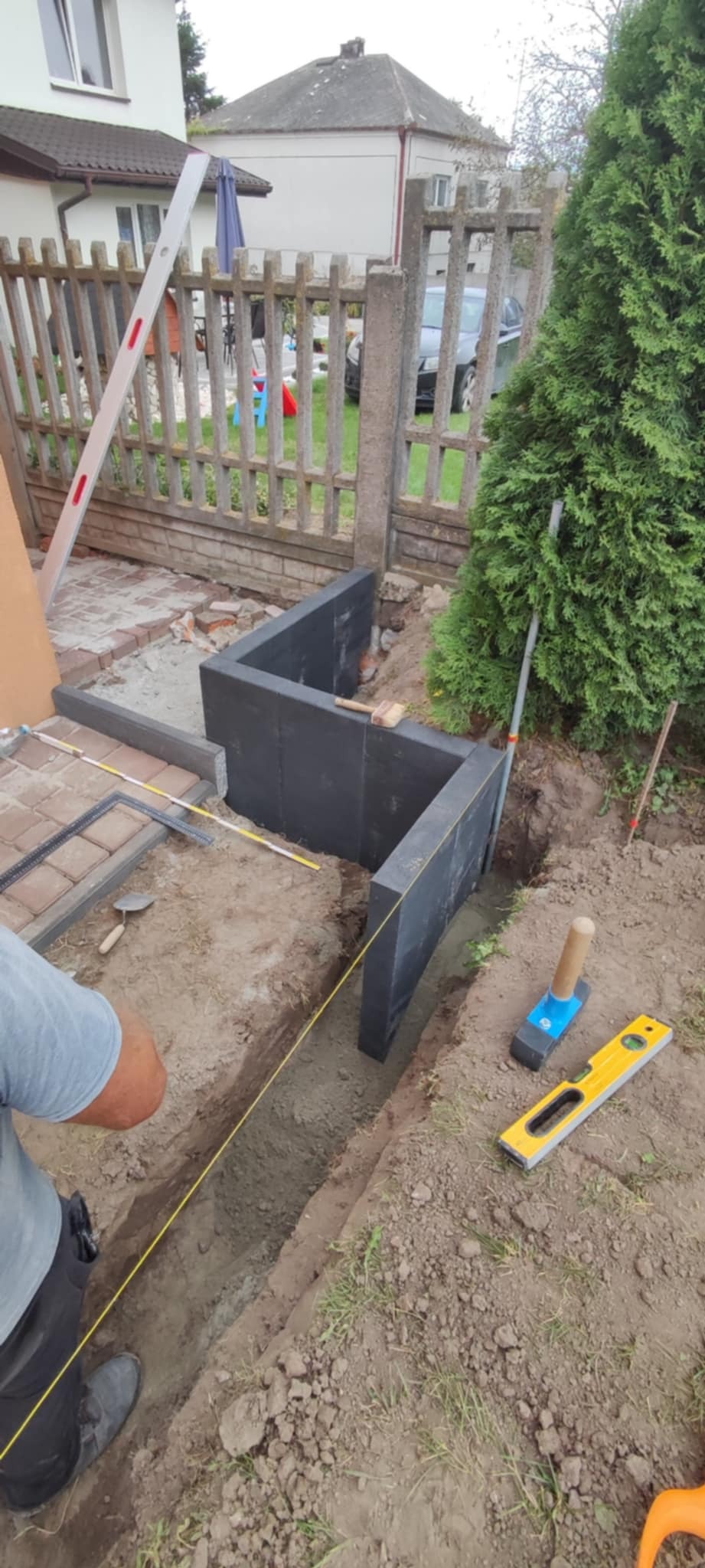 Wykop pod fundament z ustawionymi szalunkami z czarnych płyt, wytyczony sznurkiem, obok leżą poziomica i gumowy młotek, w tle widoczny częściowo ułożony bruk i ogrodzenie.