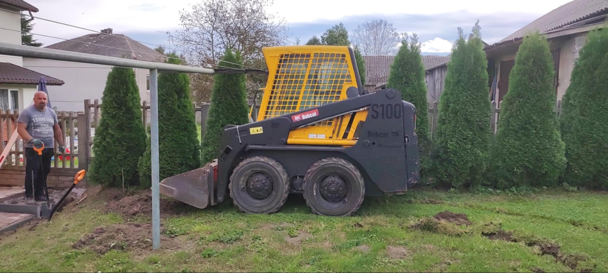 Żółto-czarna miniładowarka Bobcat S100 na trawniku, obok pracownik z łopatą i fragmenty kostki brukowej, widoczne tuje i domy w tle.